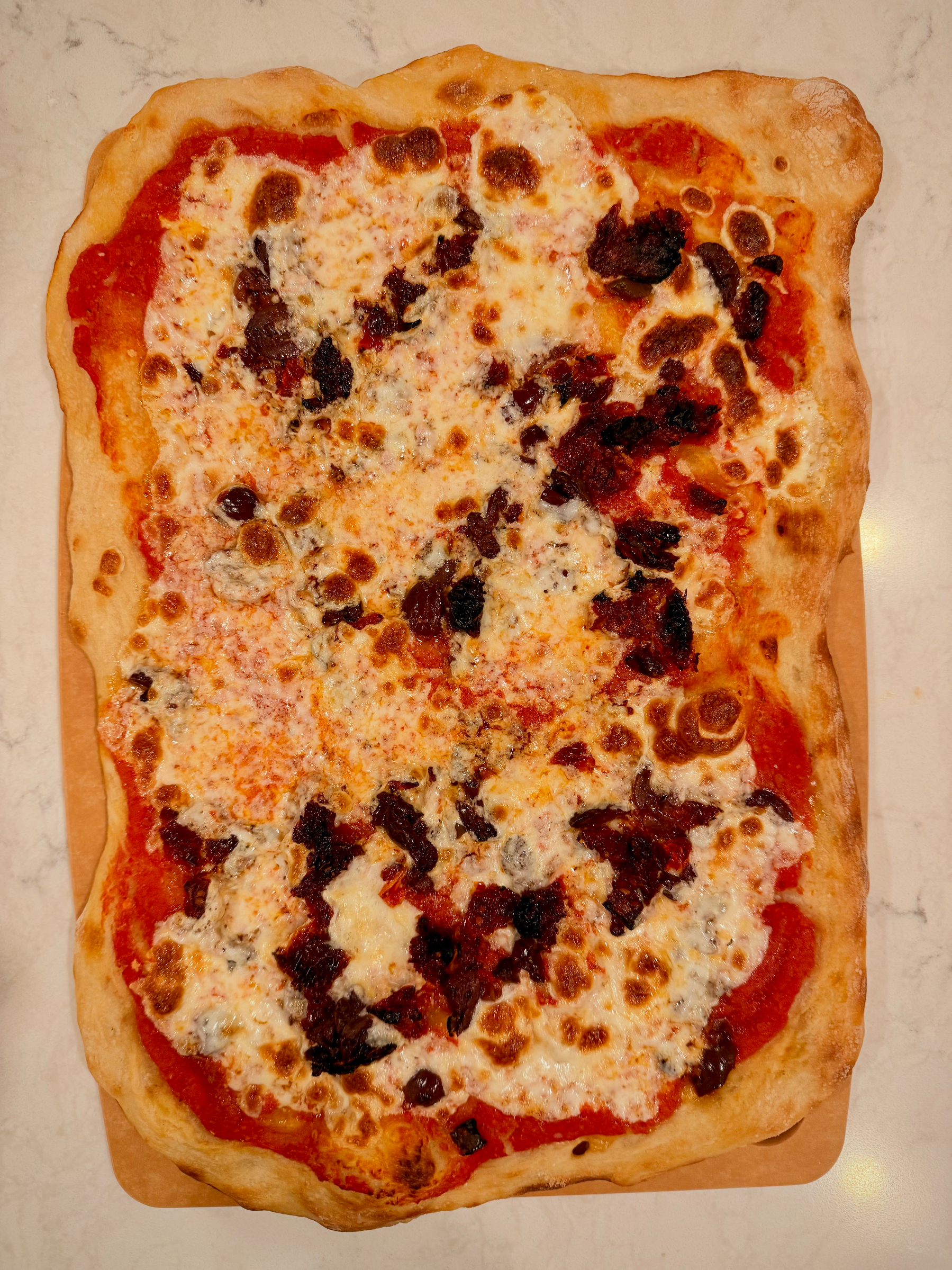 A rectangular pizza with a golden-brown crust is topped with melted cheese, tomato sauce, and scattered pieces of what appears to be sun-dried tomatoes and olives.