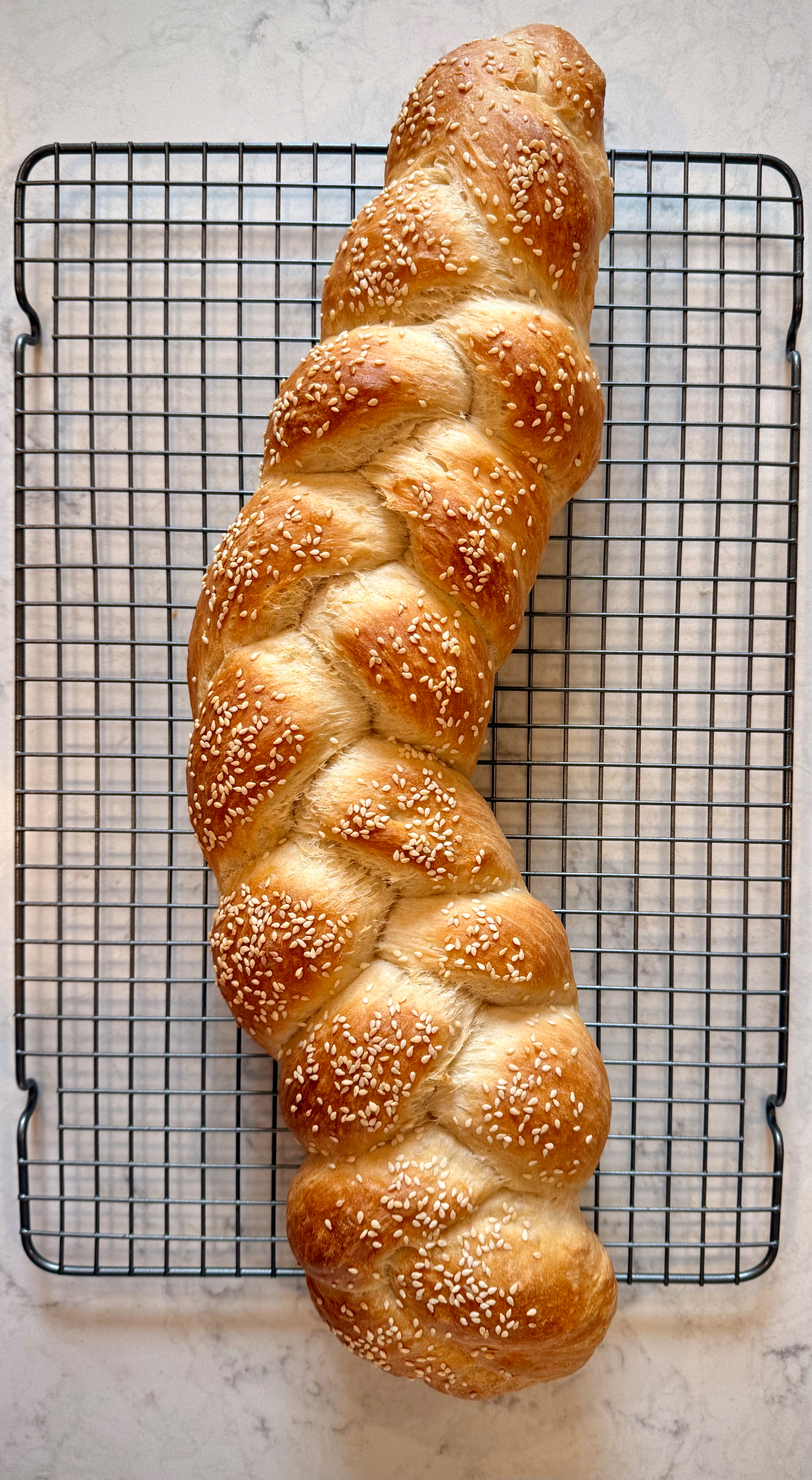 A freshly baked braided loaf topped with sesame seeds rests on a cooling rack.