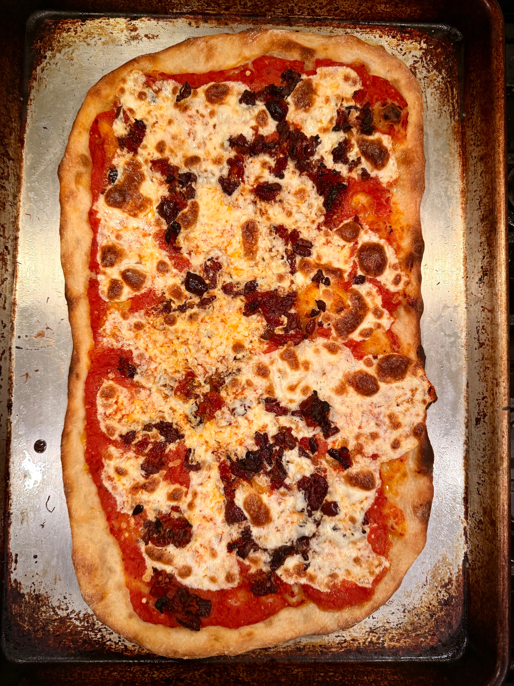 A rectangular pizza with a crispy crust, topped with melted cheese and scattered pieces of sun-dried tomatoes and olives, is placed on a baking sheet.