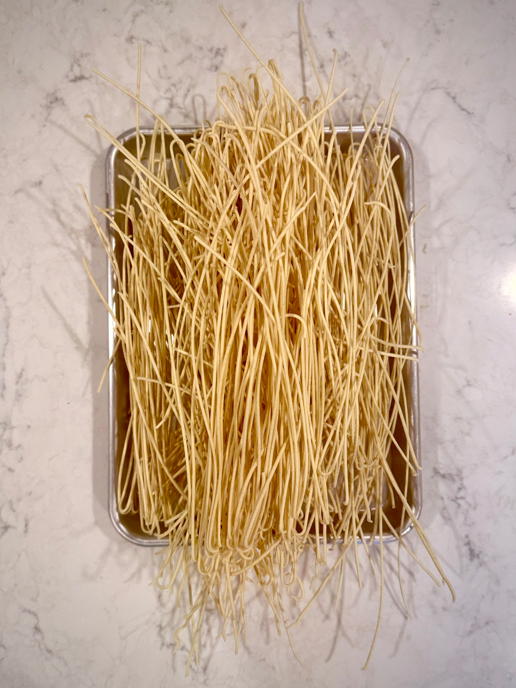 A baking tray is filled with long, uncooked spaghetti noodles arranged on a marble countertop.