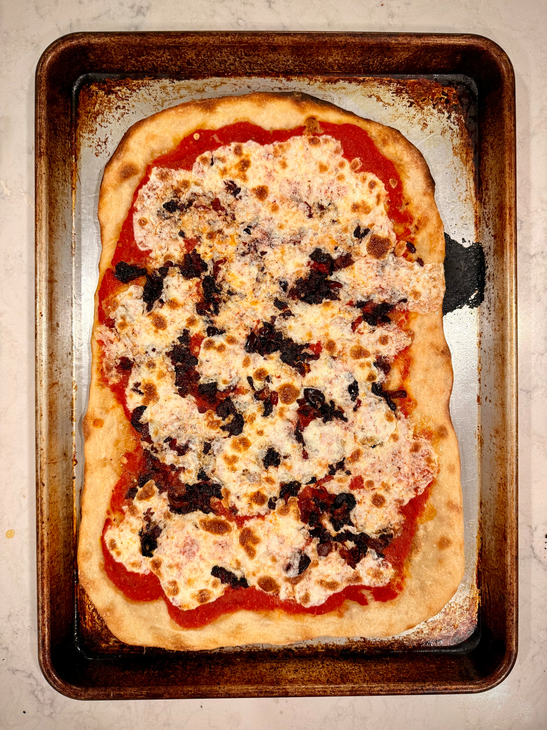 A rectangular pizza with melted cheese and toppings like olives on a baking sheet.