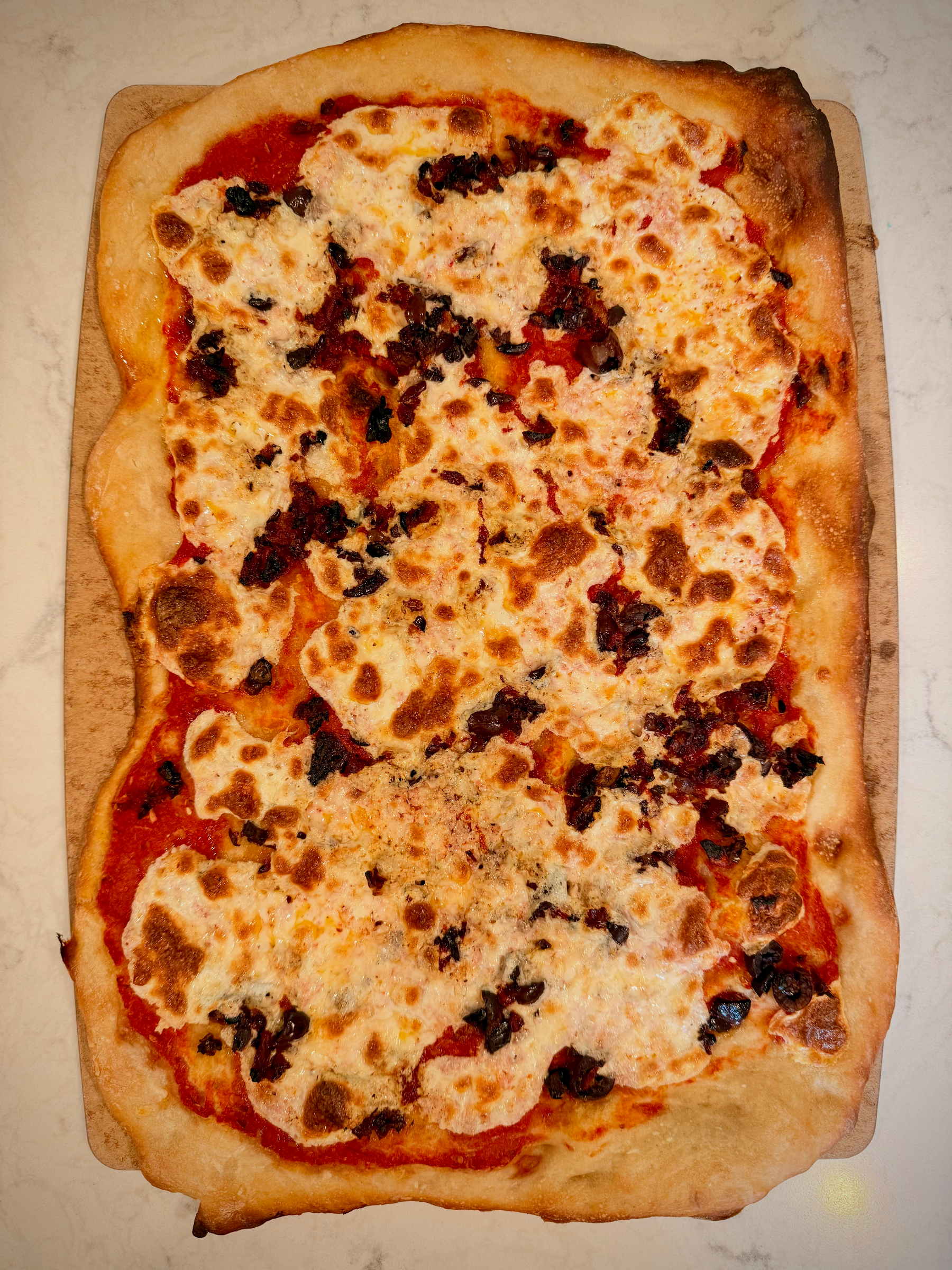 A rectangular pizza with melted cheese and dark toppings, on a wooden peel.