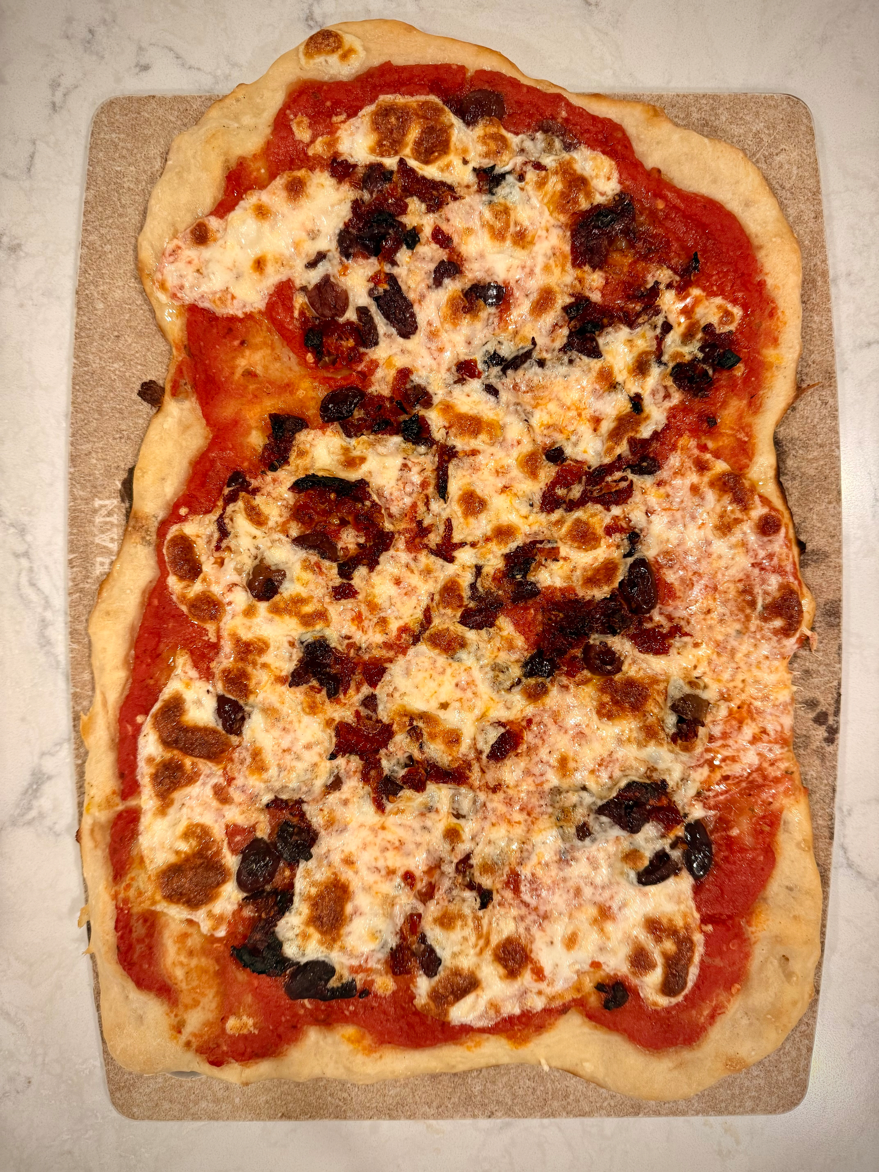 A homemade rectangular pizza topped with melted cheese and scattered pieces of black olives sits on a baking stone.