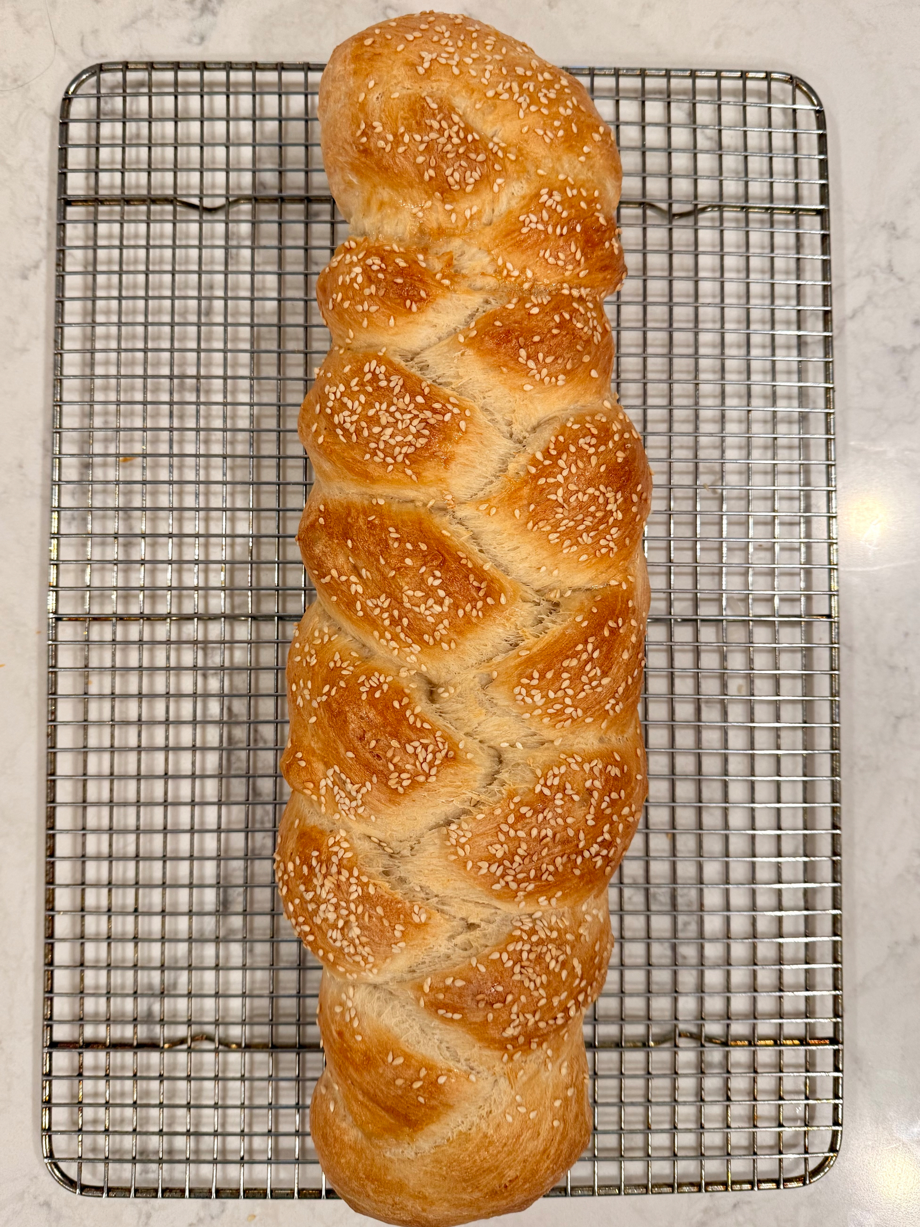A freshly baked, golden brown braided loaf topped with sesame seeds rests on a cooling rack.