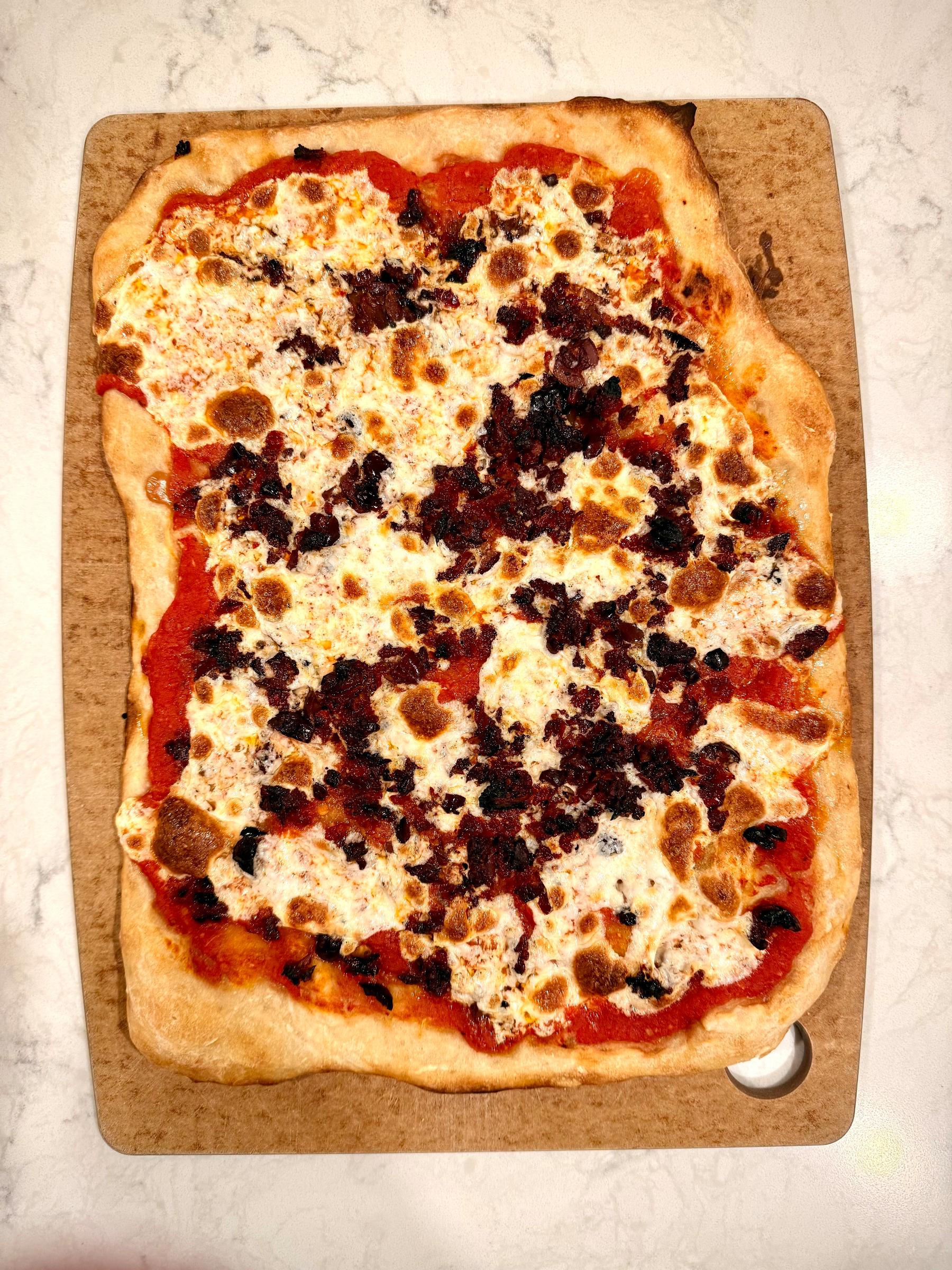 A rectangular pizza topped with cheese, tomato sauce, and pieces of meat or vegetables rests on a wooden board.
