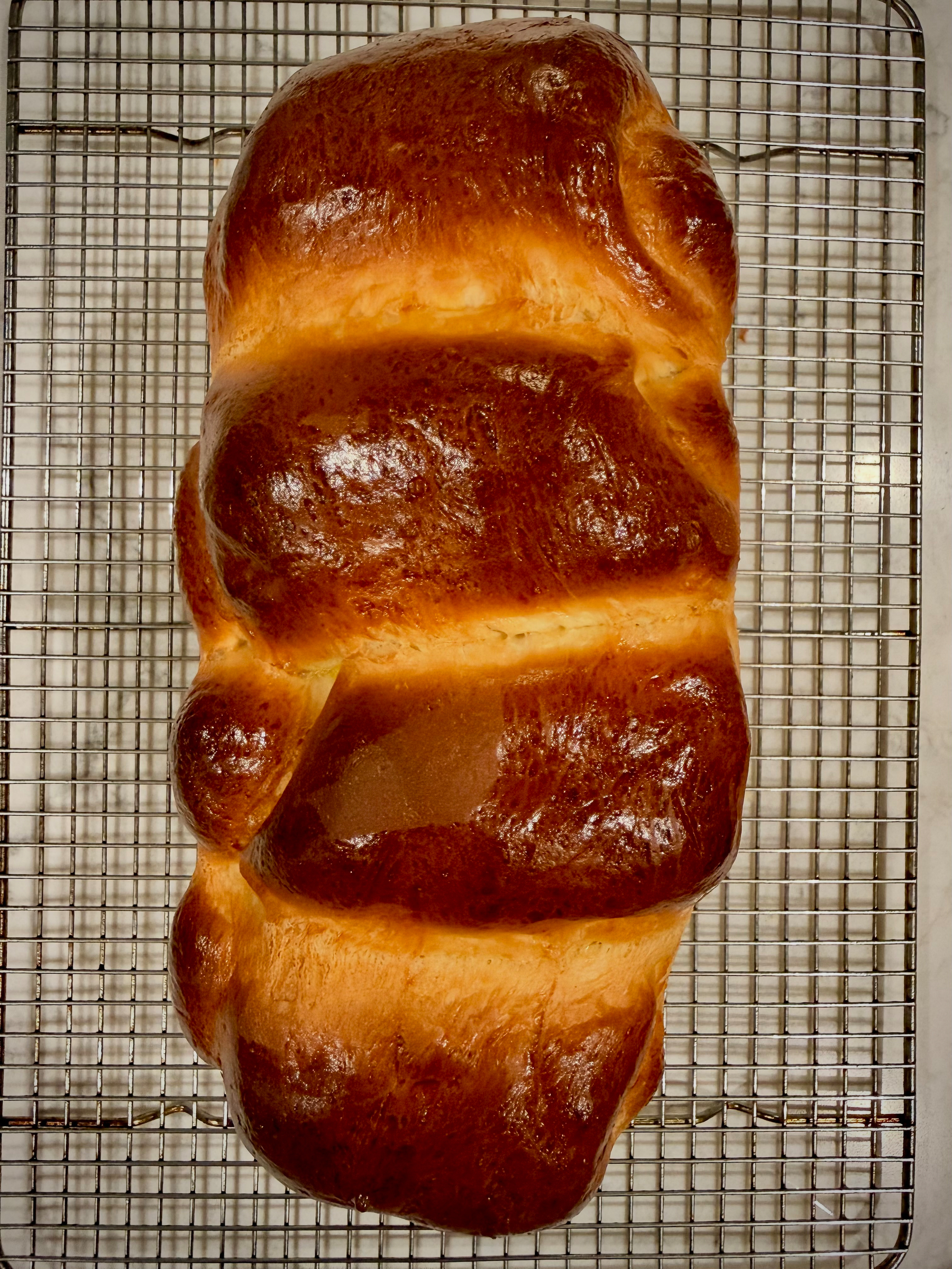 A freshly baked loaf of bread with a shiny, golden-brown crust is resting on a metal cooling rack.