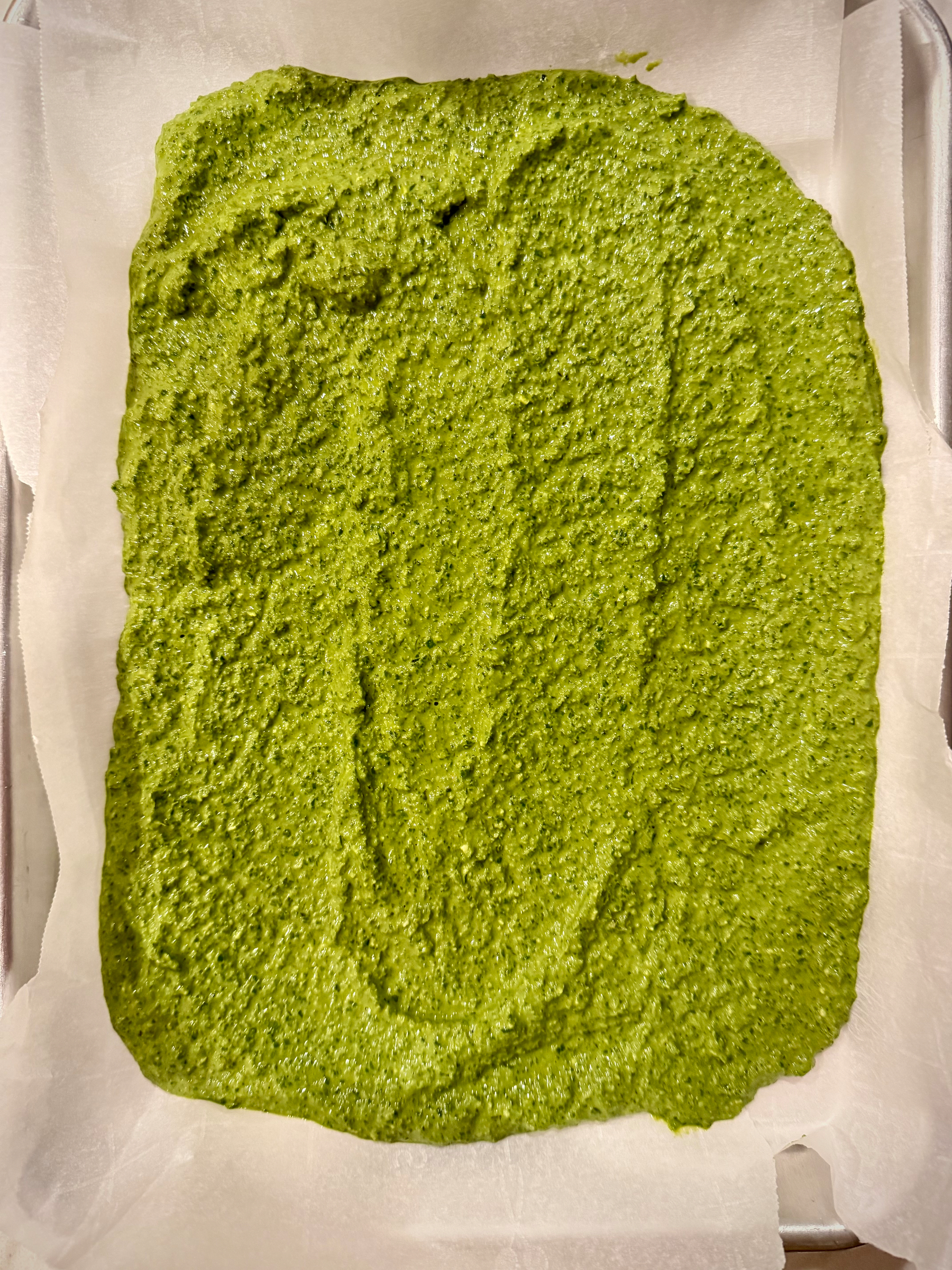 A green, textured paste or mixture is spread evenly on a rectangular sheet of parchment paper.