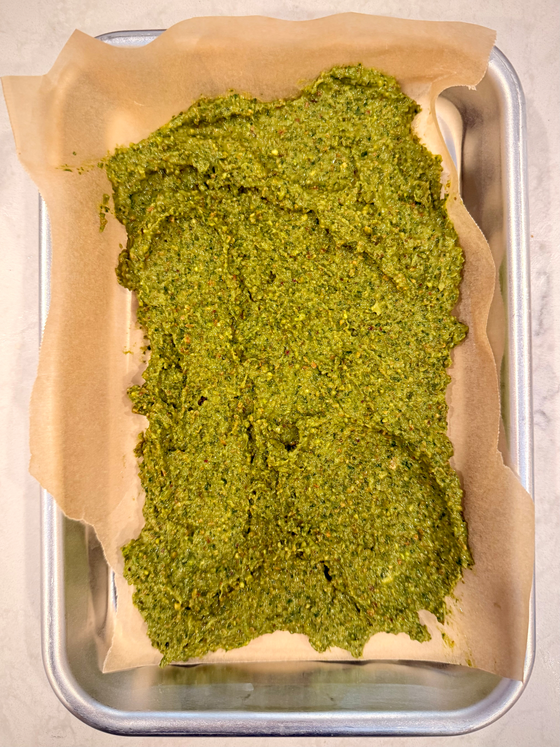 A rectangular sheet pan holds a layer of green pesto spread on parchment paper.
