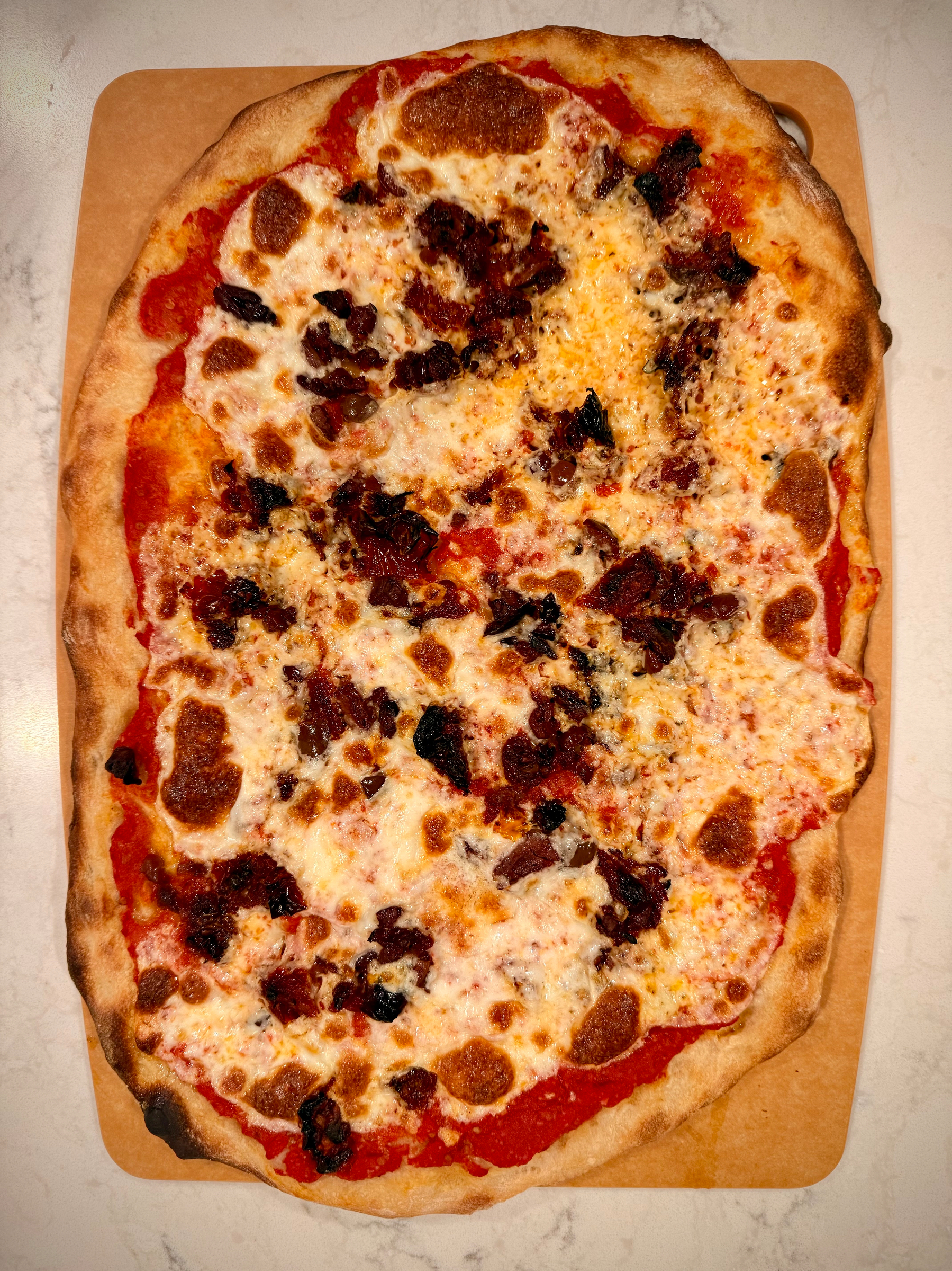 A rectangular pizza with a crispy crust topped with melted cheese, tomato sauce, and scattered pieces of olives and sun-dried tomatoes .