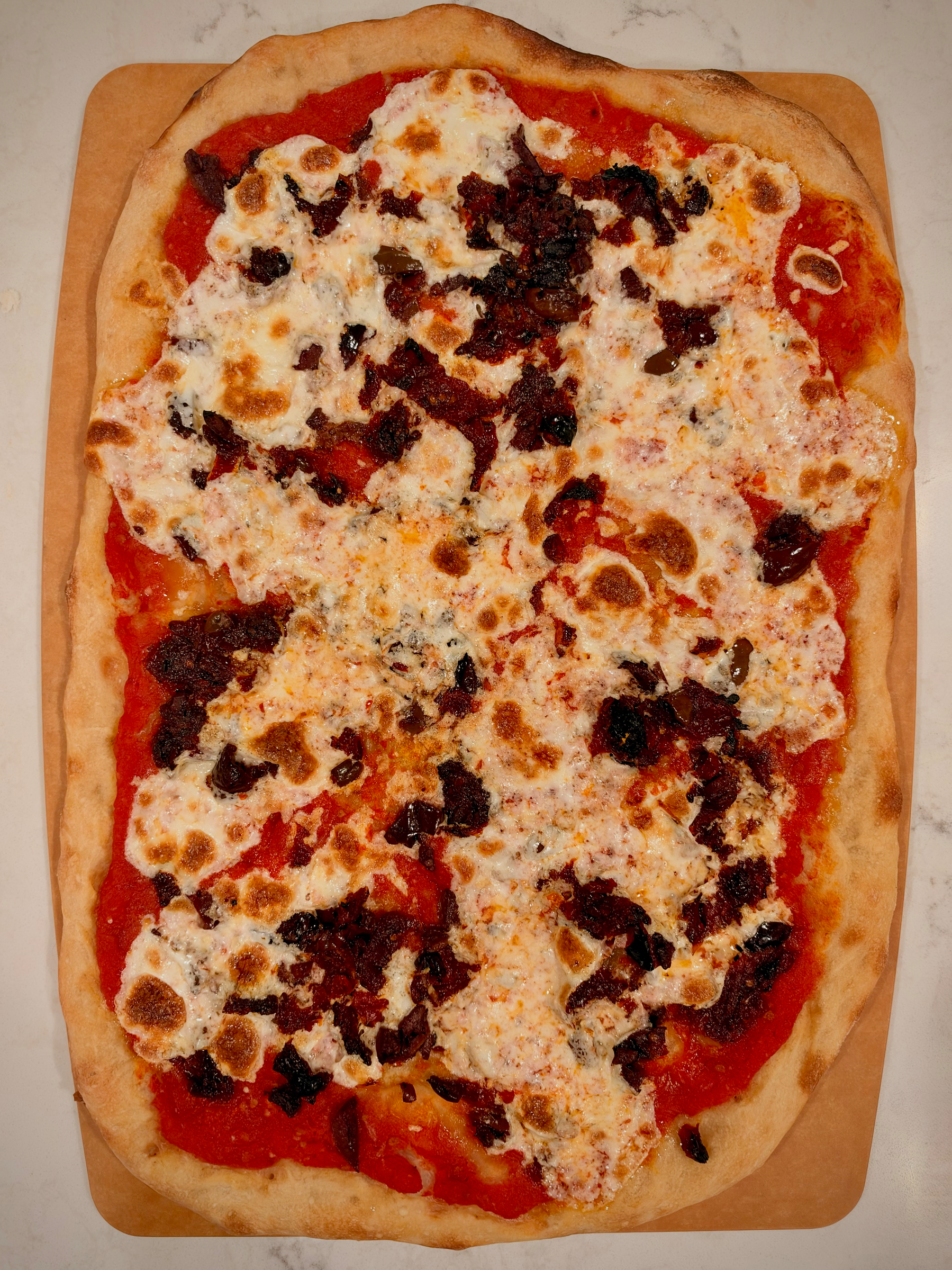 A rectangular pizza topped with melted cheese, tomato sauce, and scattered pieces of dark-colored toppings sits on a wooden board.