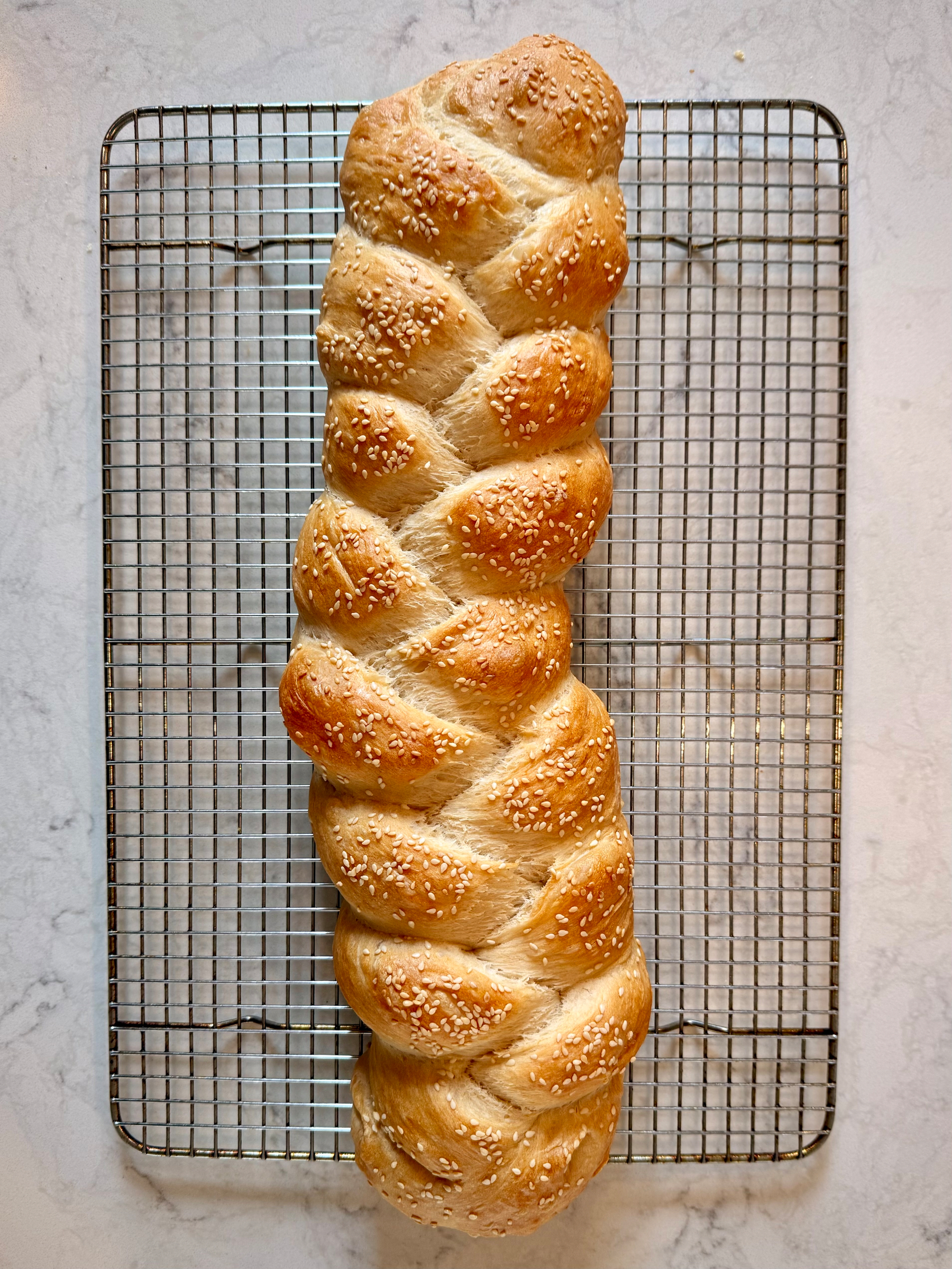 A loaf of braided bread topped with sesame seeds is cooling on a wire rack placed on a marble surface.