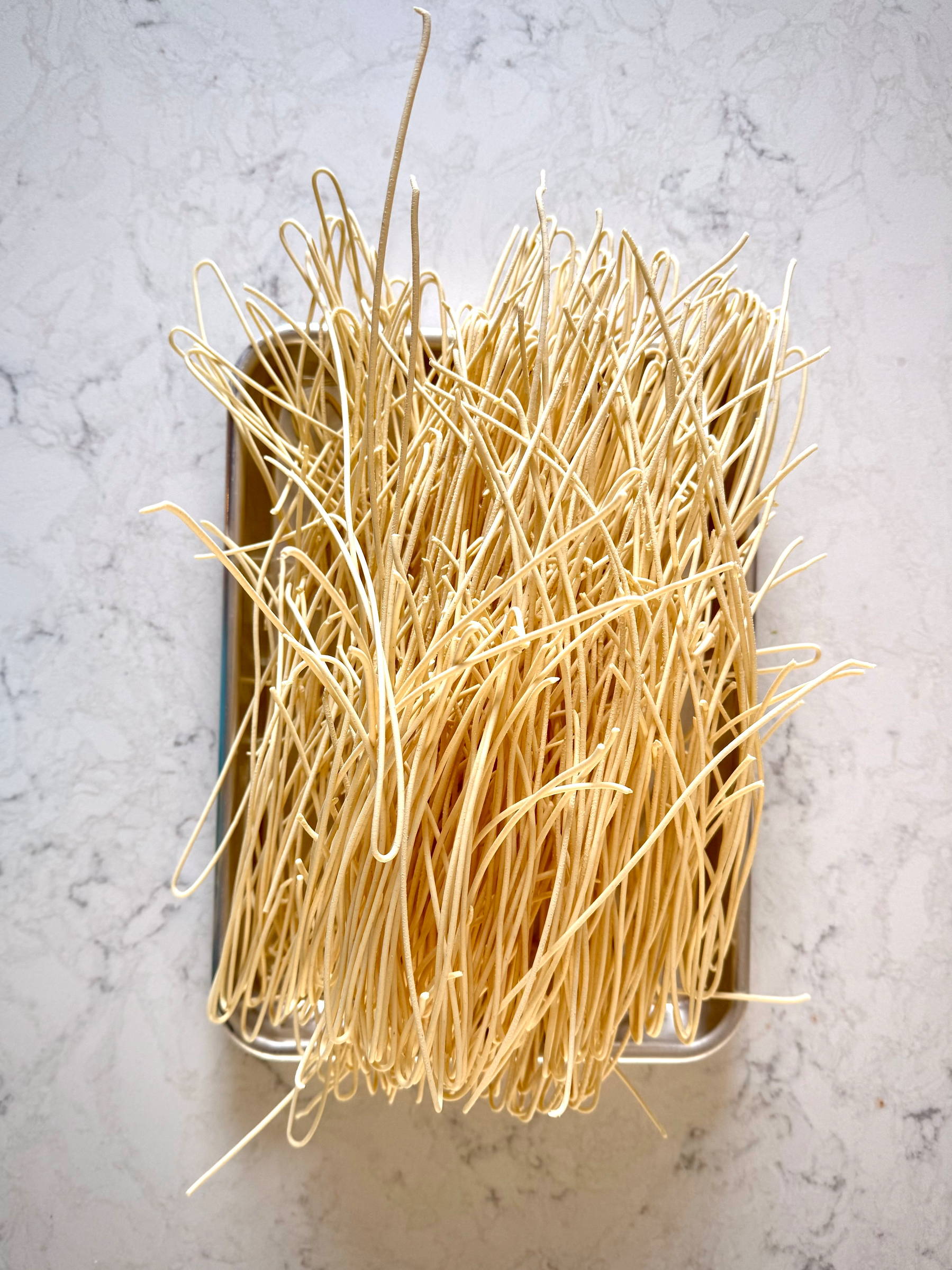 A top-down view of long, thin strands of fresh pasta neatly draped and piled in a rectangular metal tray. The pale yellow noodles overlap and loop irregularly, filling the tray and spilling slightly over the edges. The tray rests on a light, marble-patterned countertop, giving the scene a clean, homemade pasta-making feel.
