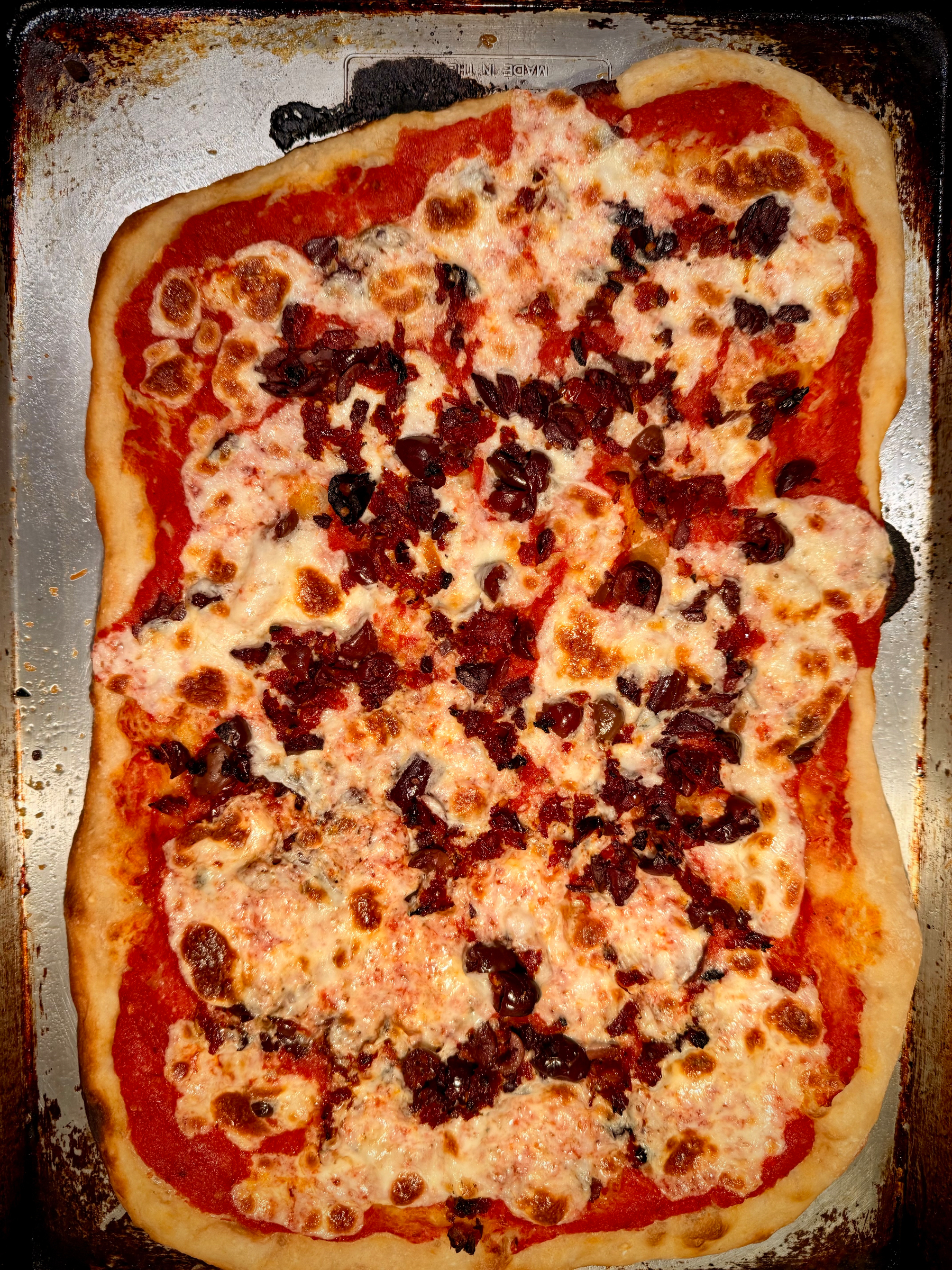 This picture shows a rectangular pizza on a baking sheet, topped with melted cheese and what appears to be olives or a similar sun dried tomato topping.