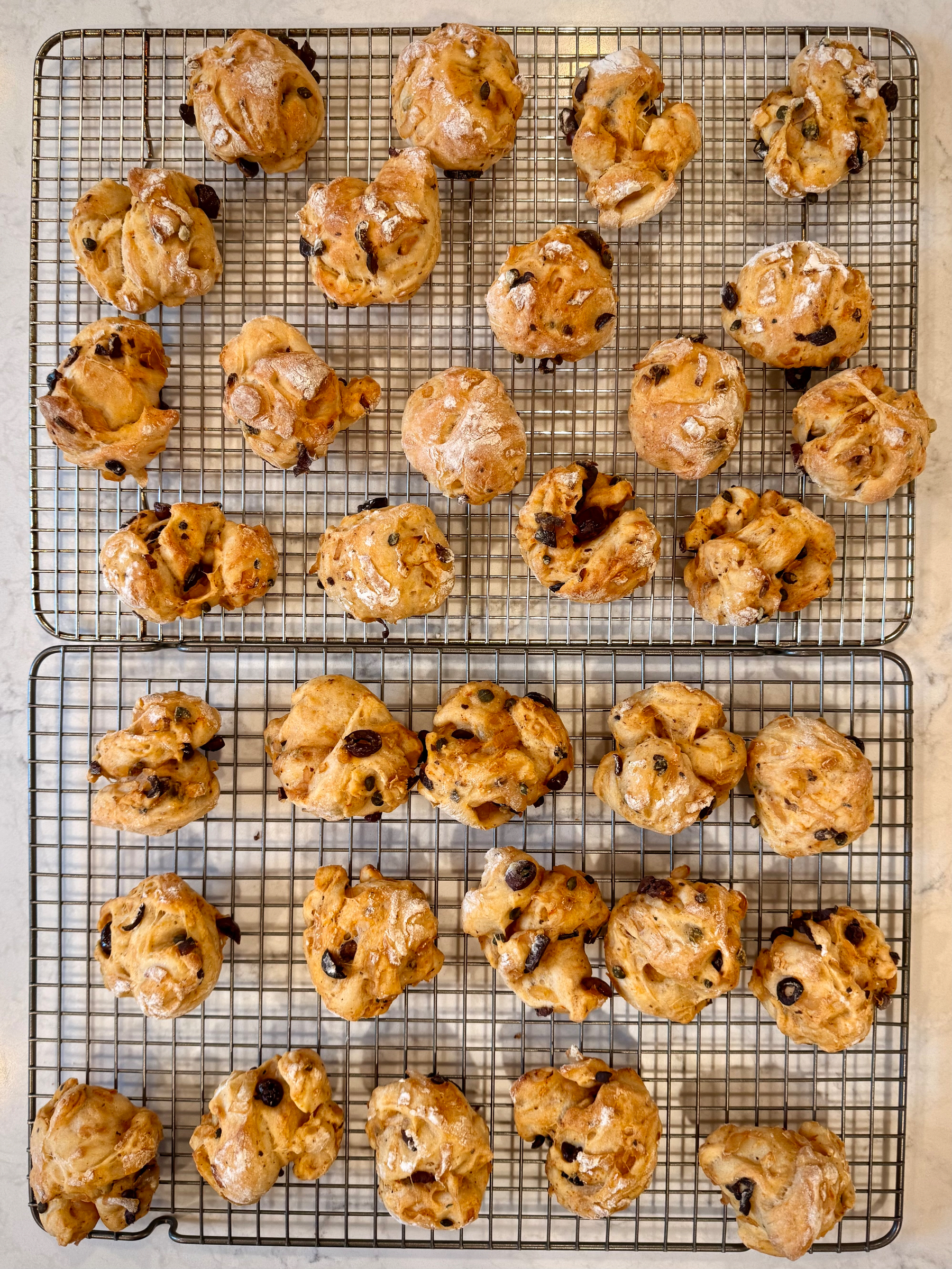 Freshly baked, golden brown rolls with olives, onions, and capers, cooling on wire racks.