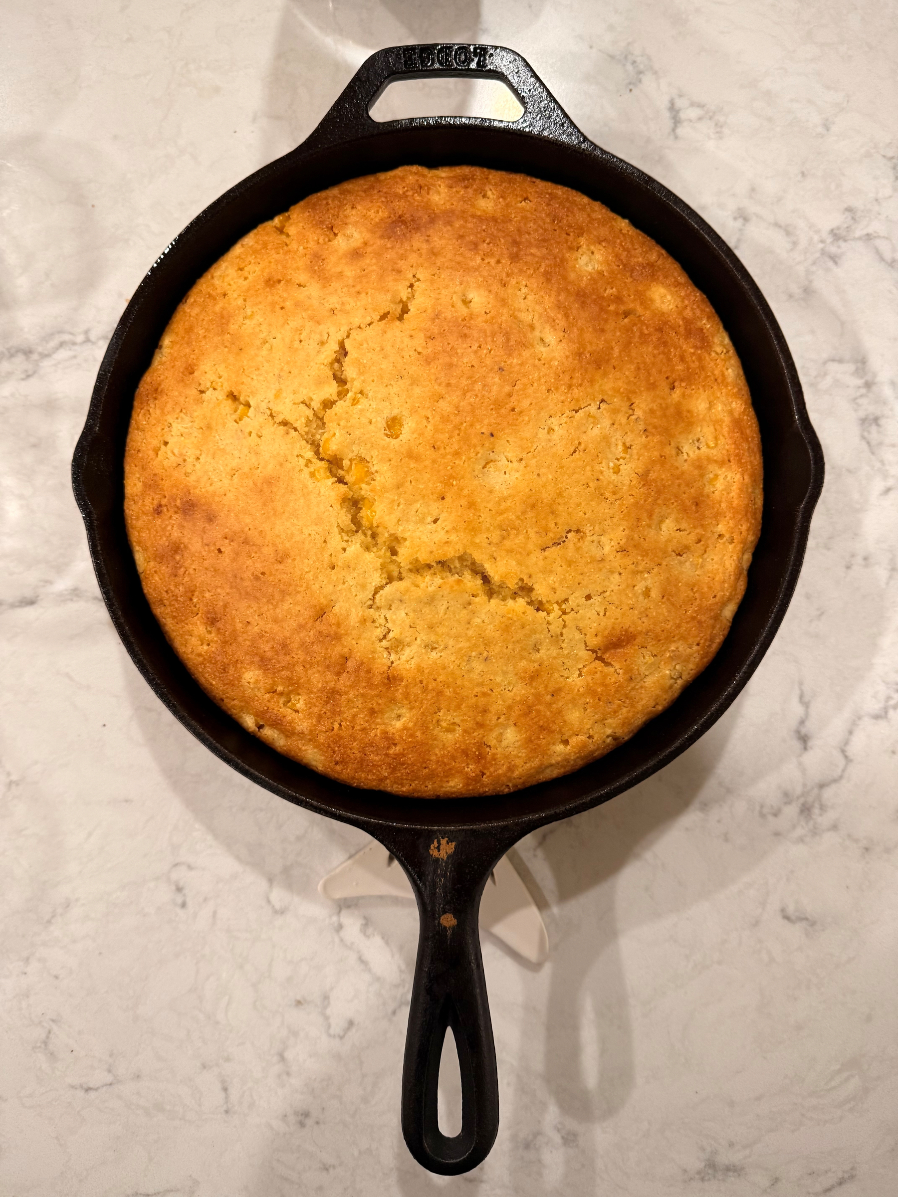 A freshly baked golden-brown cornbread is resting in a black cast iron skillet on a marble surface.