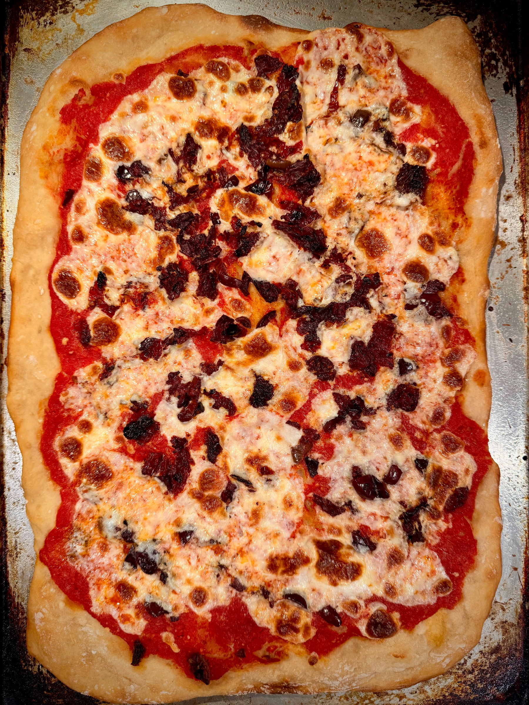 A rectangular homemade pizza with melted cheese, tomato sauce, and pieces of tomato and olive on a baking sheet.