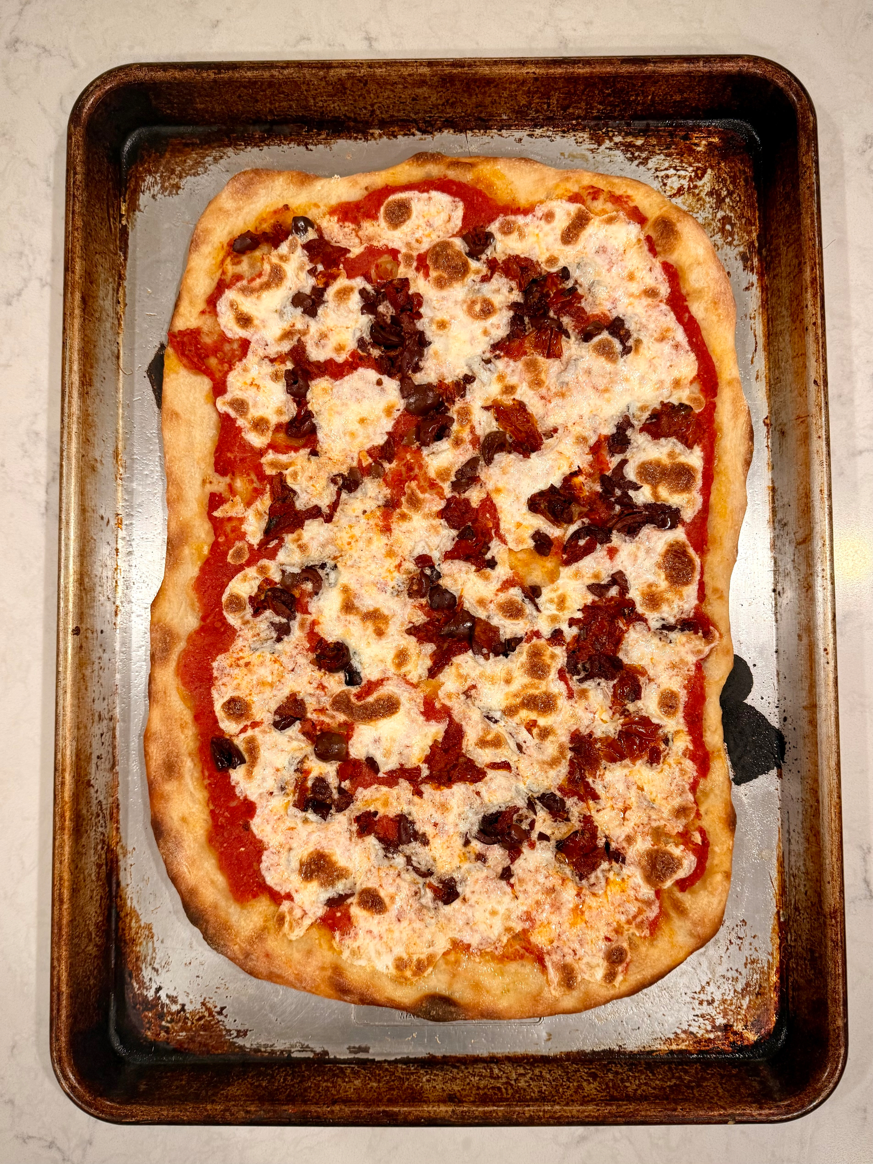 A rectangular homemade pizza topped with melted cheese and pieces of tomato and olive rests on a baking sheet.