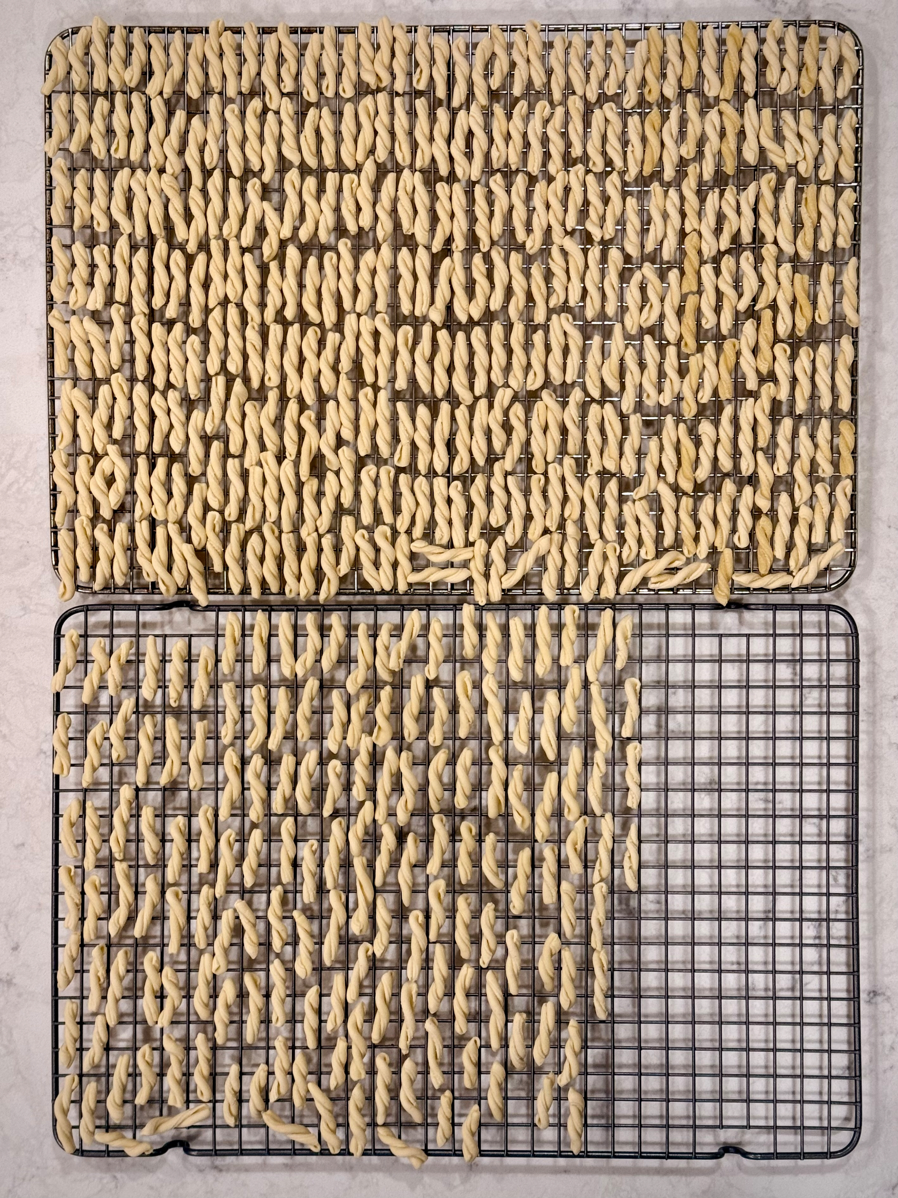 A large quantity of twist-like pasta is drying on two stacked wire racks.