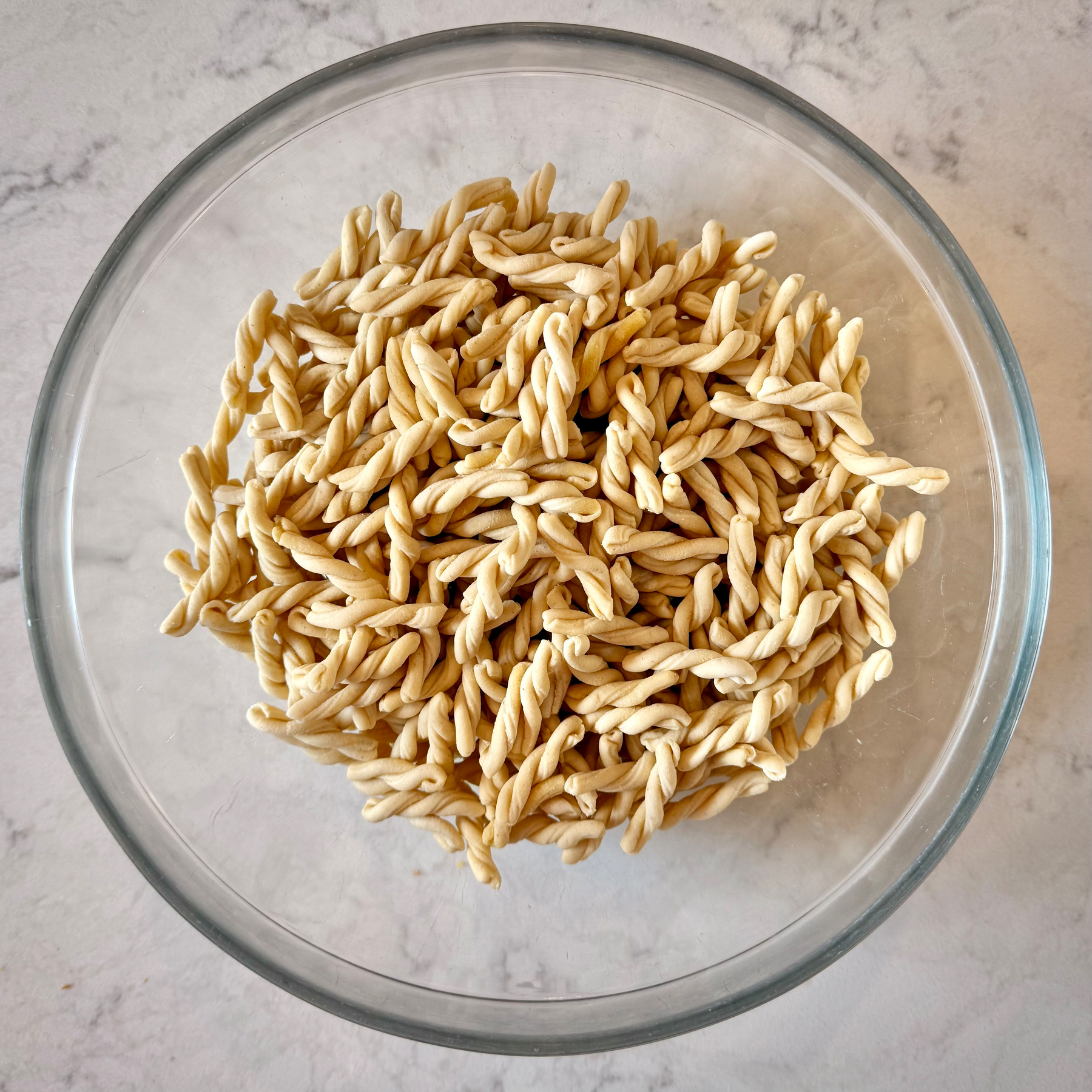 A glass bowl is filled with uncooked twisted pasta on a marble surface.
