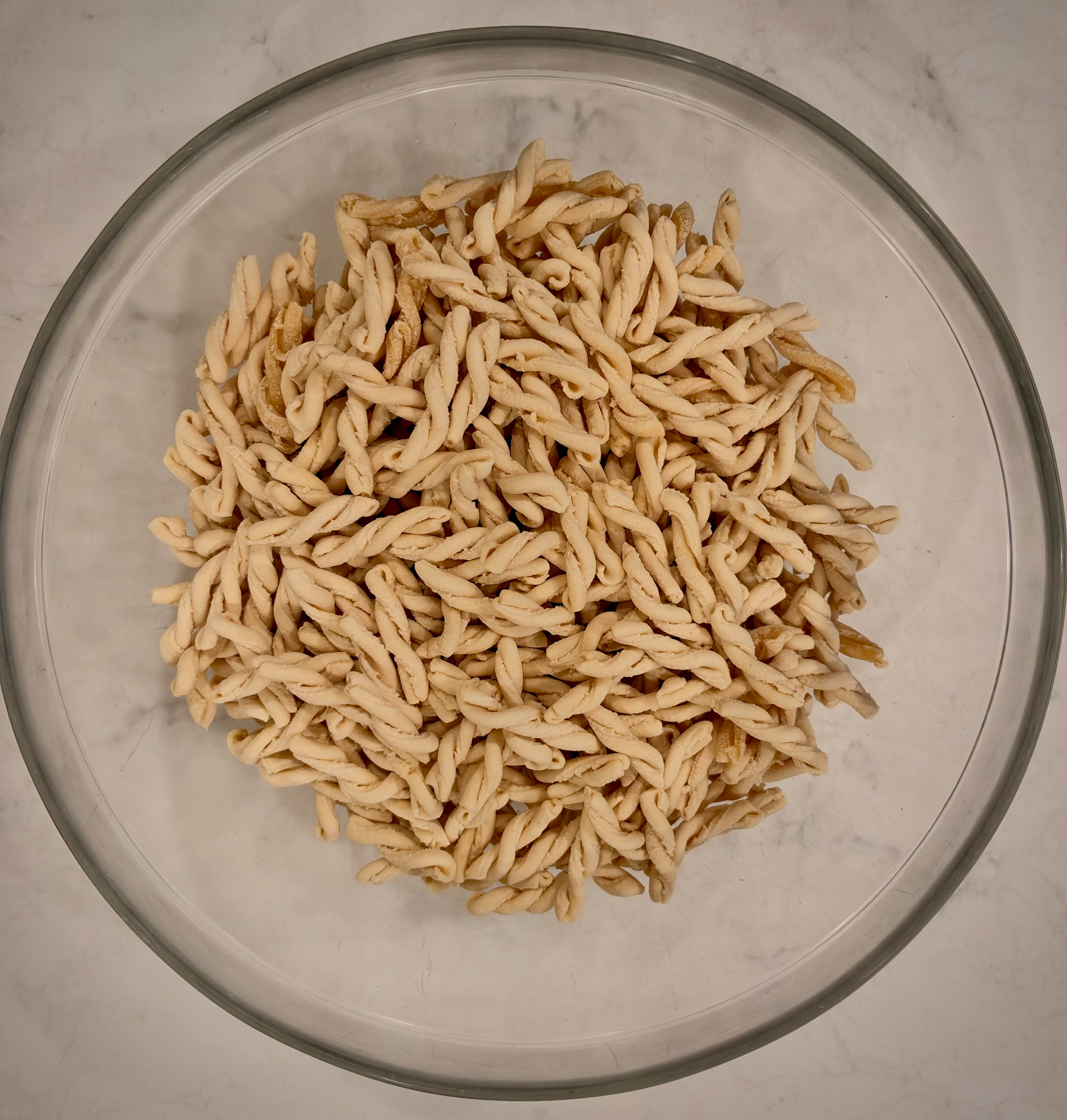A clear glass bowl is filled with a large amount of twisted, uncooked pasta on a light-colored surface.