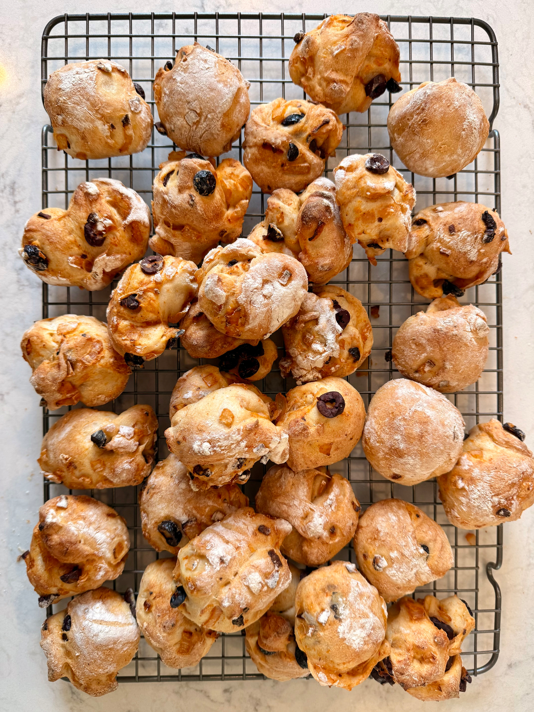A cooling rack holds frsshly baked, golden brown buns dotted with olives and onions.