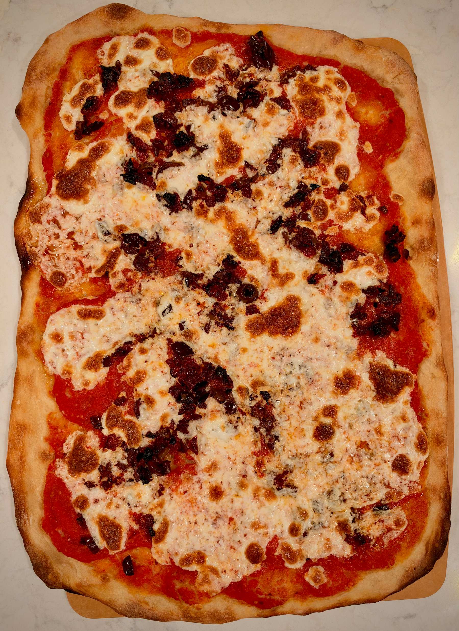 A rectangular pizza topped with melted cheese, tomato sauce, and small pieces of dark toppings is placed on a light-colored surface.