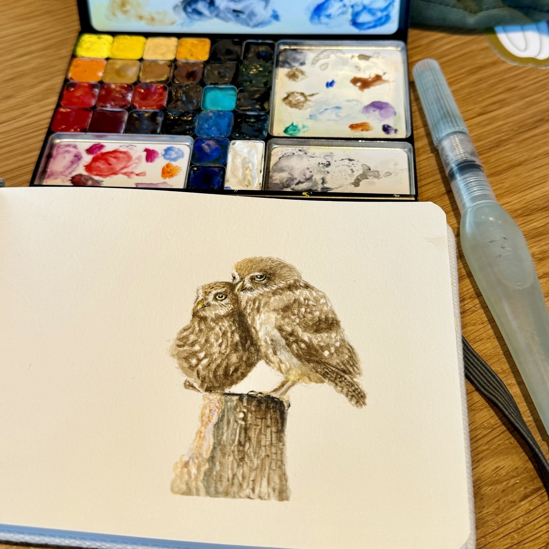 A watercolor of a couple of owls in a sketchbook with a palette and water brush in the background. 