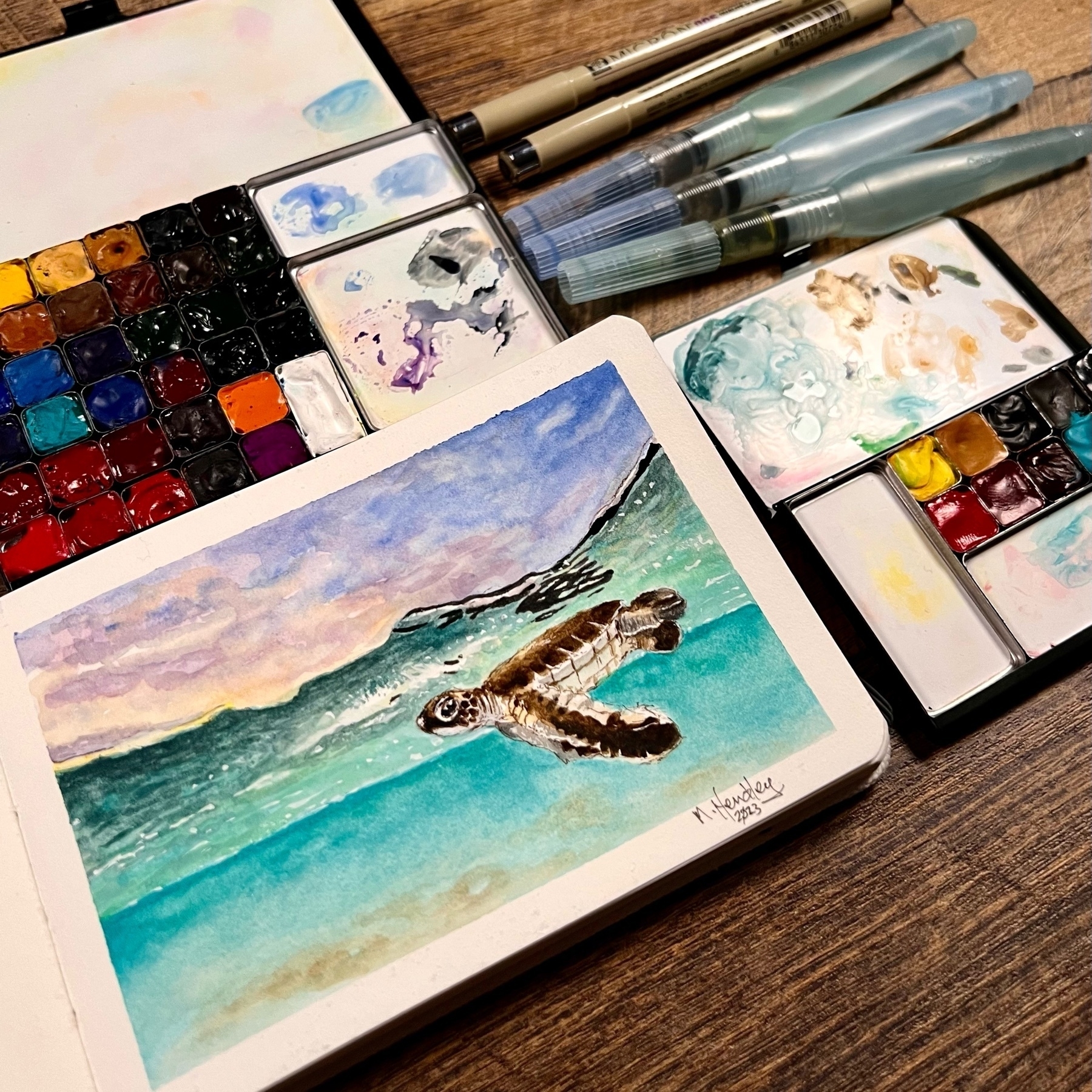 a watercolor painting of a baby sea turtle just below the surface on the ocean under a purplish sky. the book is on a table with two art palettes, brushes and pens. 