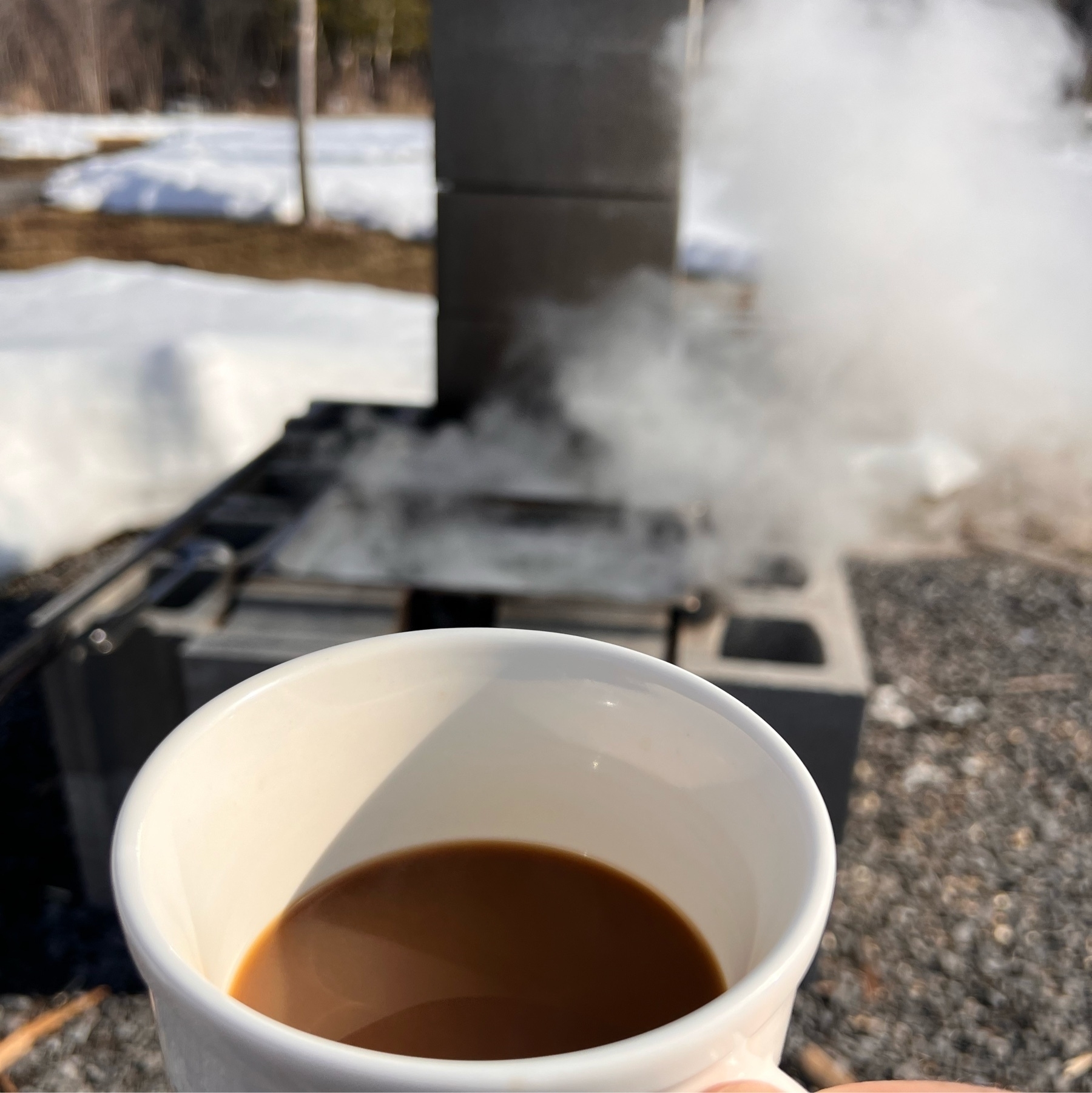 cup of coffee held in hand with a homemade maple sap boiler built with cinder blocks in the background. 