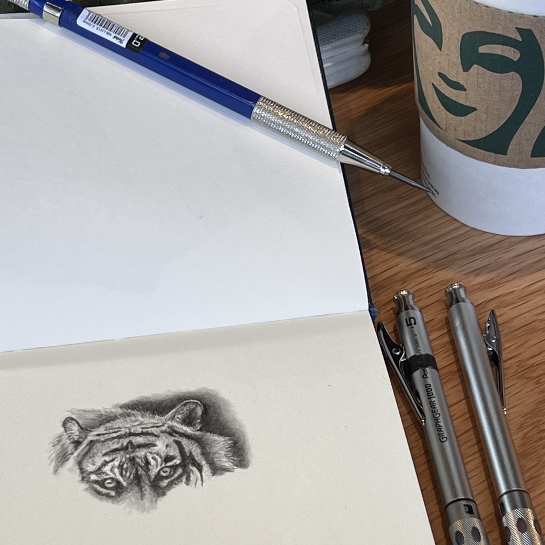 A drawing of a tiger’s head in a sketchbook. Surrounded by three mechanical pencils and a Starbucks coffee cup. 