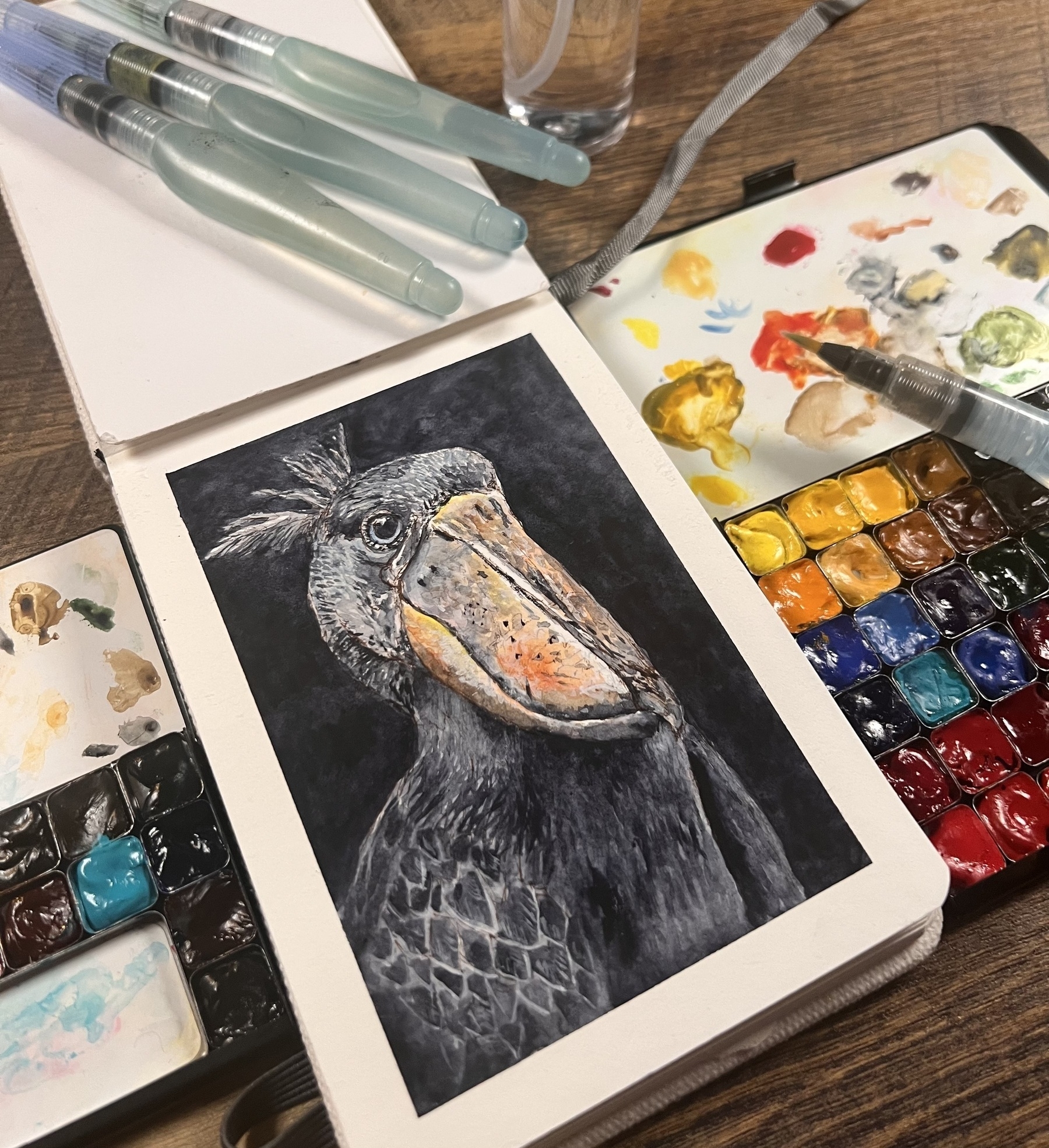 Shoebill stork watercolor in a sketchbook. Surrounded by two palettes and some water brushes 