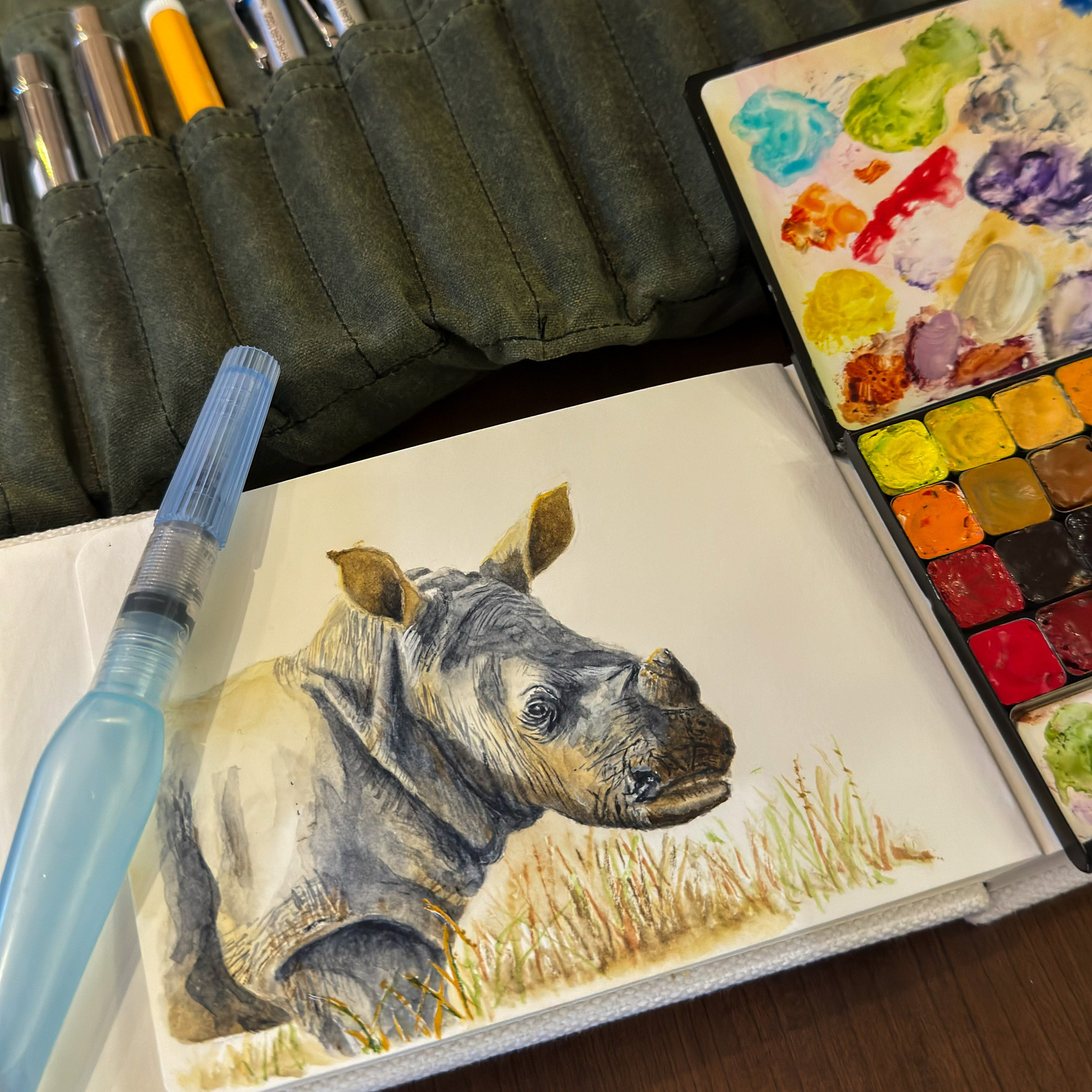 Watercolor painting of a baby rhino lying in the grass, with a palette of vibrant colors and watercolor brushes nearby.