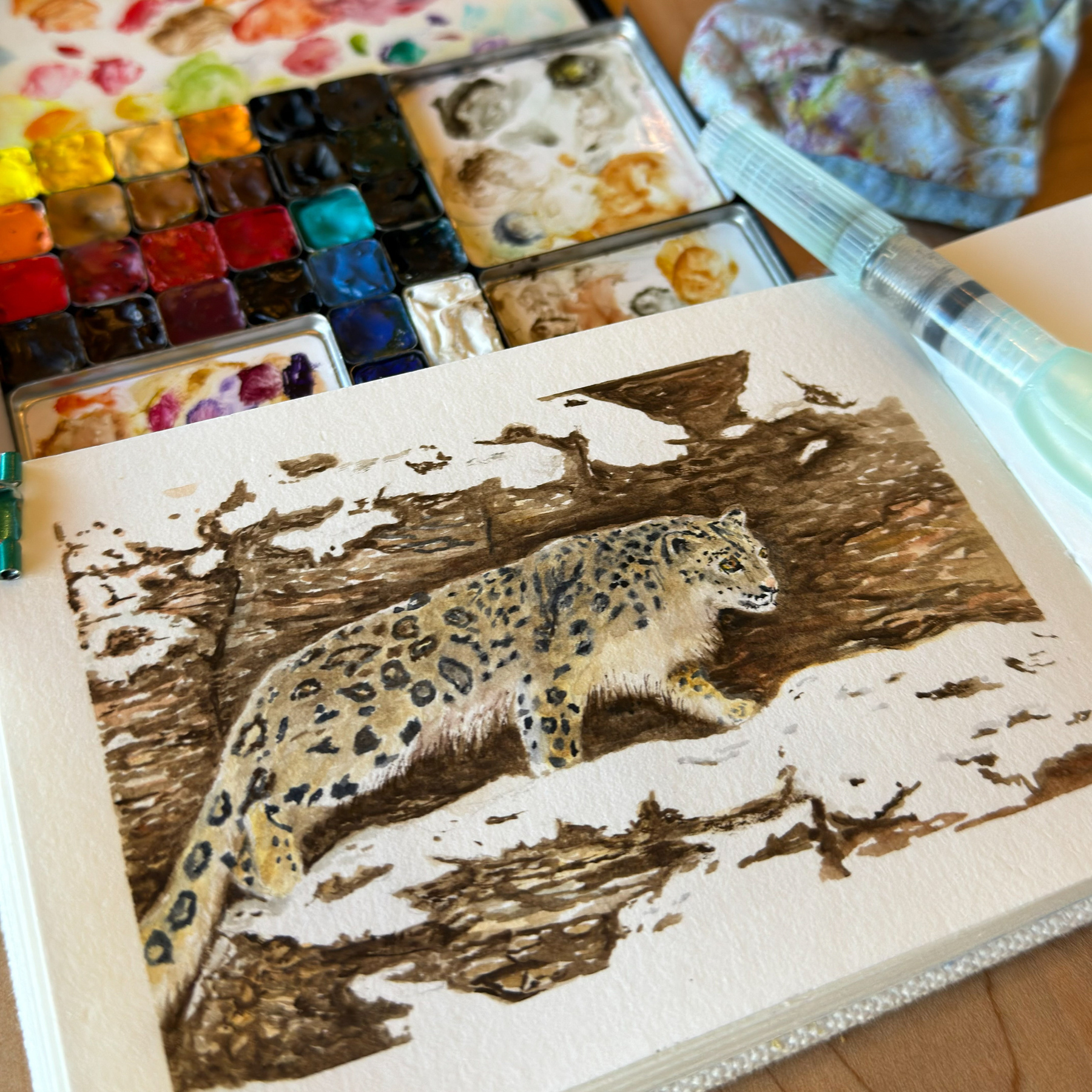 A watercolor painting in a sketchbook depicting a snow leopard with intricate spot patterns crouched on a rocky terrain, accompanied by a watercolor palette with vibrant hues and a used rag on a wooden surface.