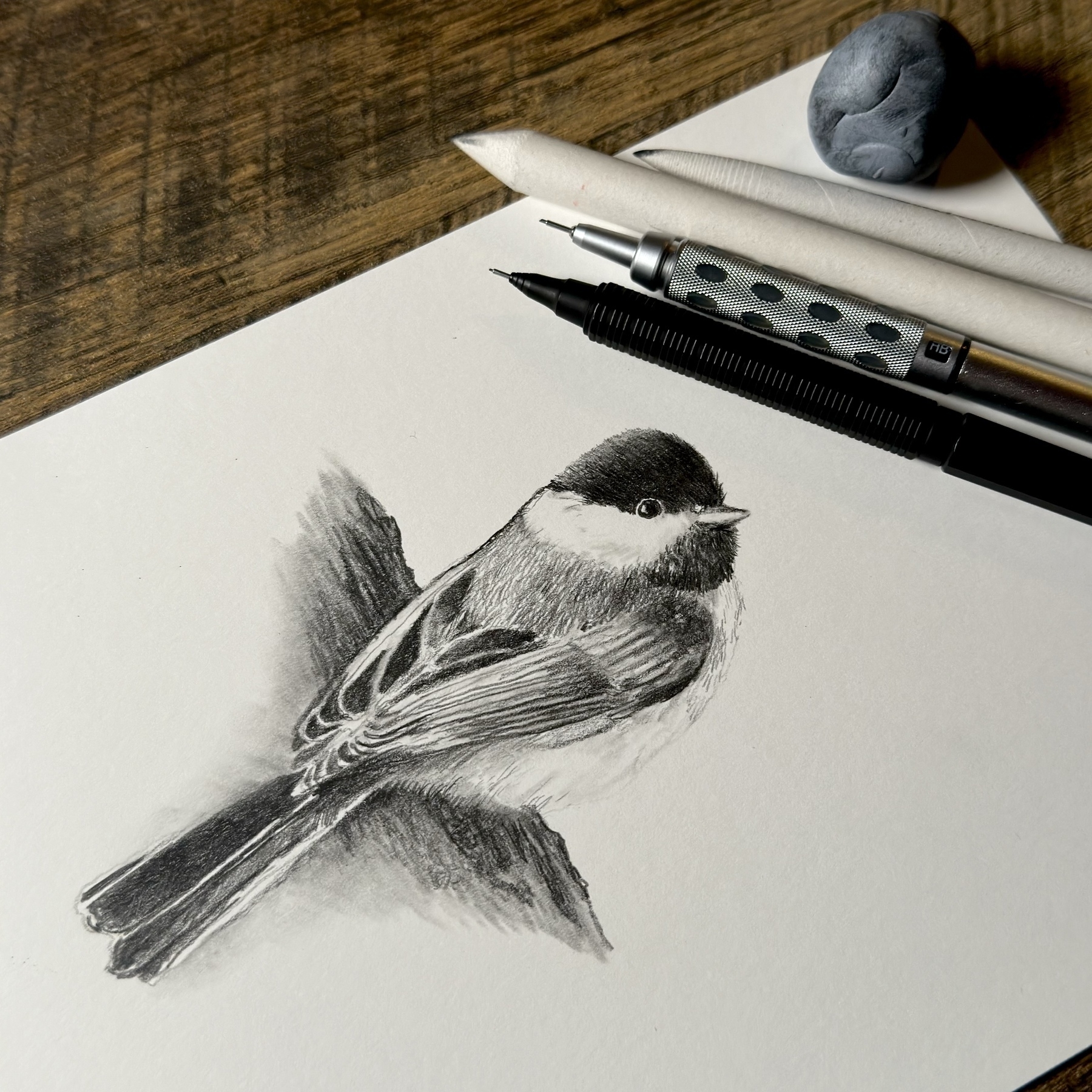 An intricate graphite pencil sketch of a black-capped chickadee positioned on the right, with its detailed feathers and distinct markings clearly visible. The sketch is on a white paper that lies on a wooden surface, surrounded by drawing tools which include a mechanical pencil, a blending stump, an eraser, and a kneaded eraser to the top left.