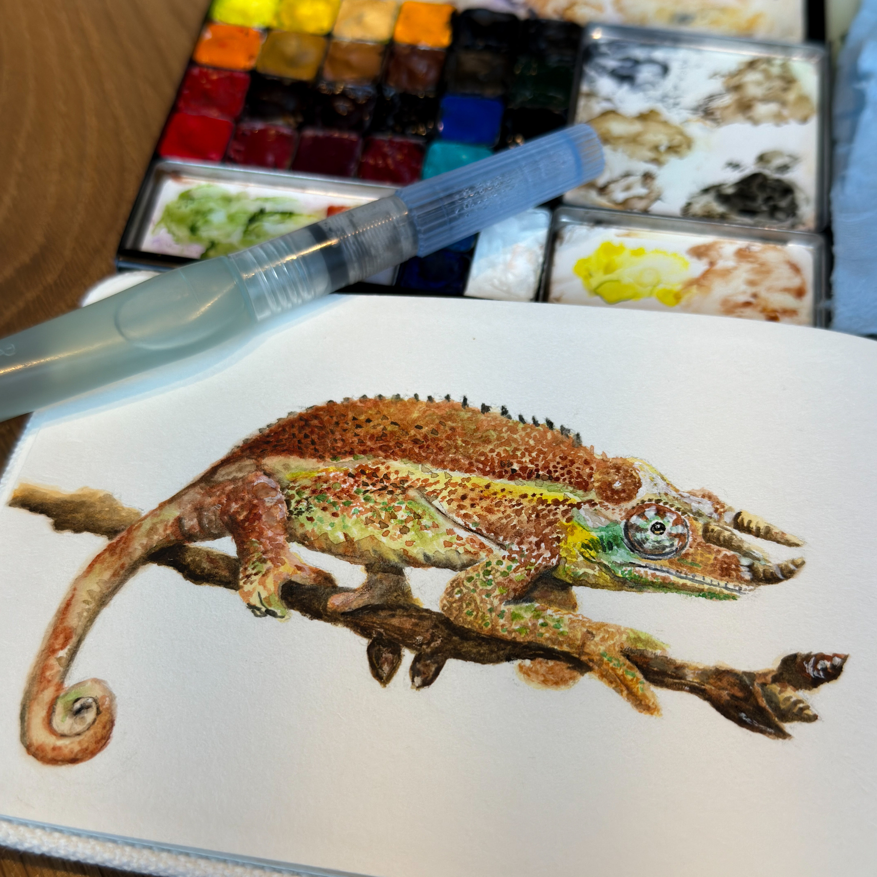 A realistic painting of a chameleon on a branch is set alongside an open watercolor palette and a filled water brush.