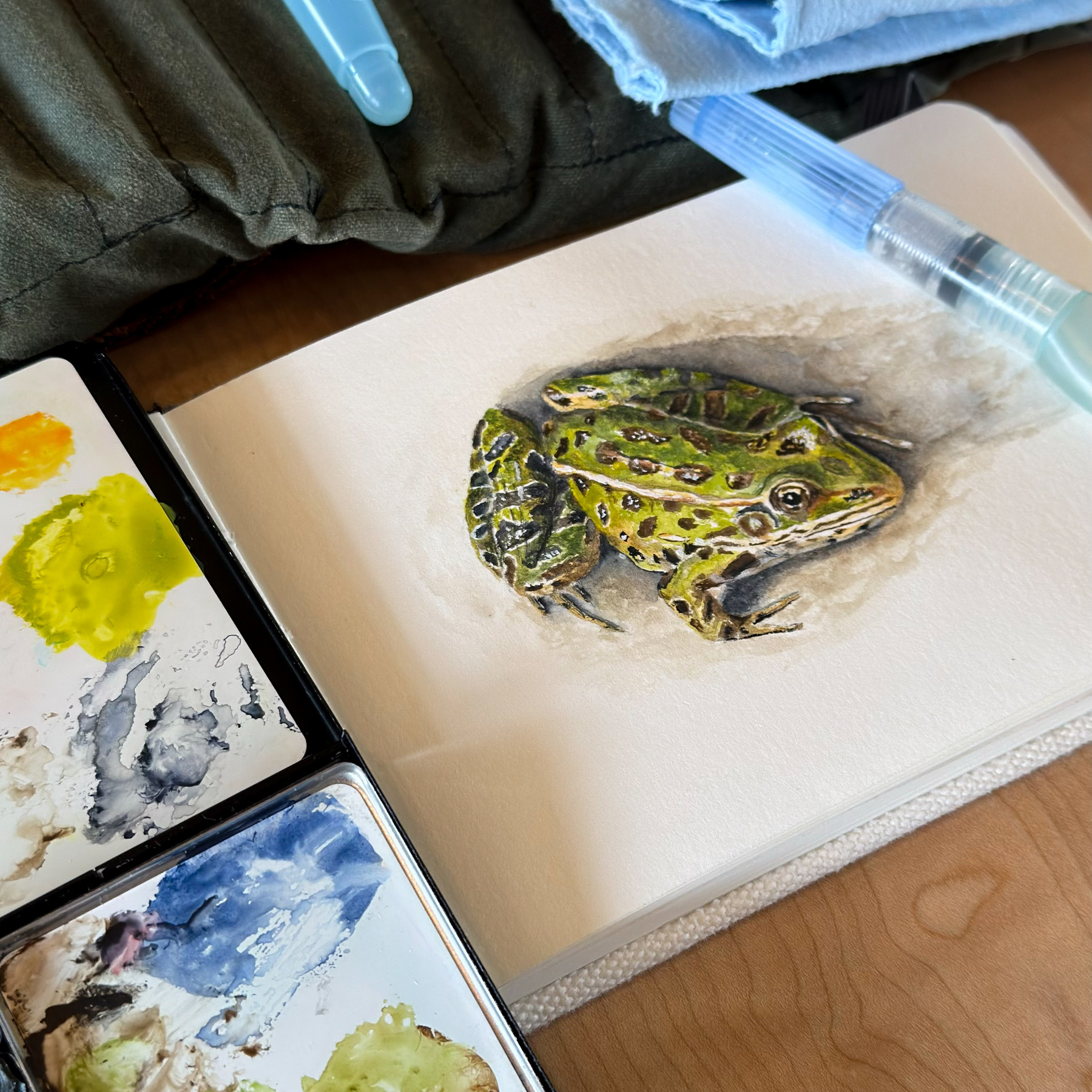 A detailed watercolor painting of a leopard frog on an open sketchbook, surrounded by watercolor paints and brushes. The green and yellow hues of the frog contrast with the earthy tones of the sketchbook and painting supplies.