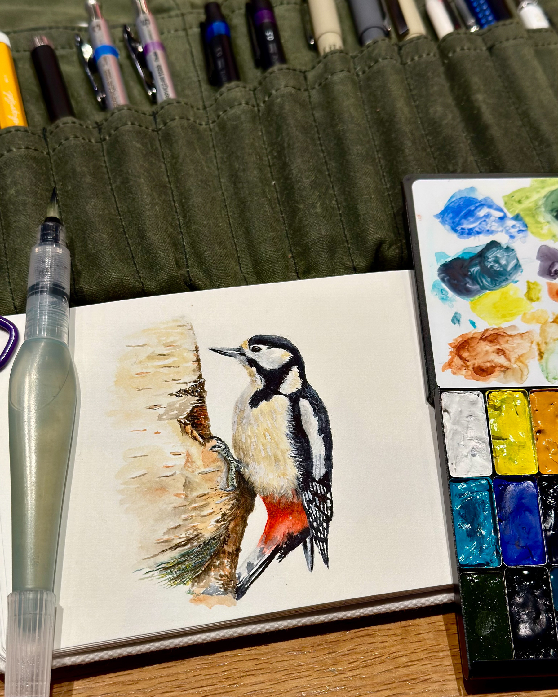 A watercolor painting of a great spotted woodpecker perched on a tree trunk, created in a sketchbook. Nearby are watercolor pans, a Pentel Aquash water brush, and an array of pens in a green fabric case.