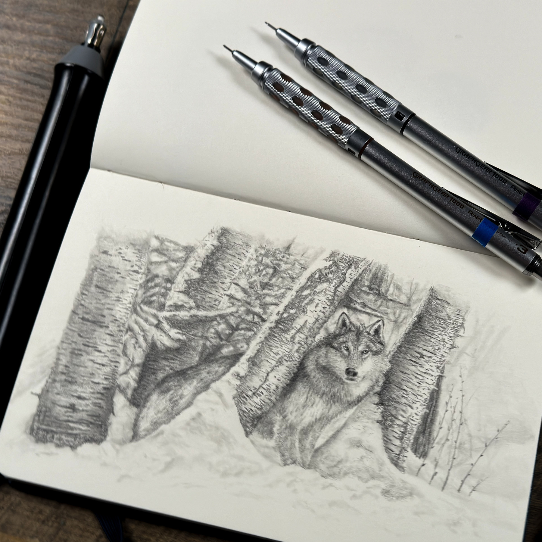 A pencil sketch of a wolf in a forest, placed on a wooden table surrounded by drawing tools including a mechanical pencil, two blenders, an electric eraser, and a piece of kneaded eraser.
