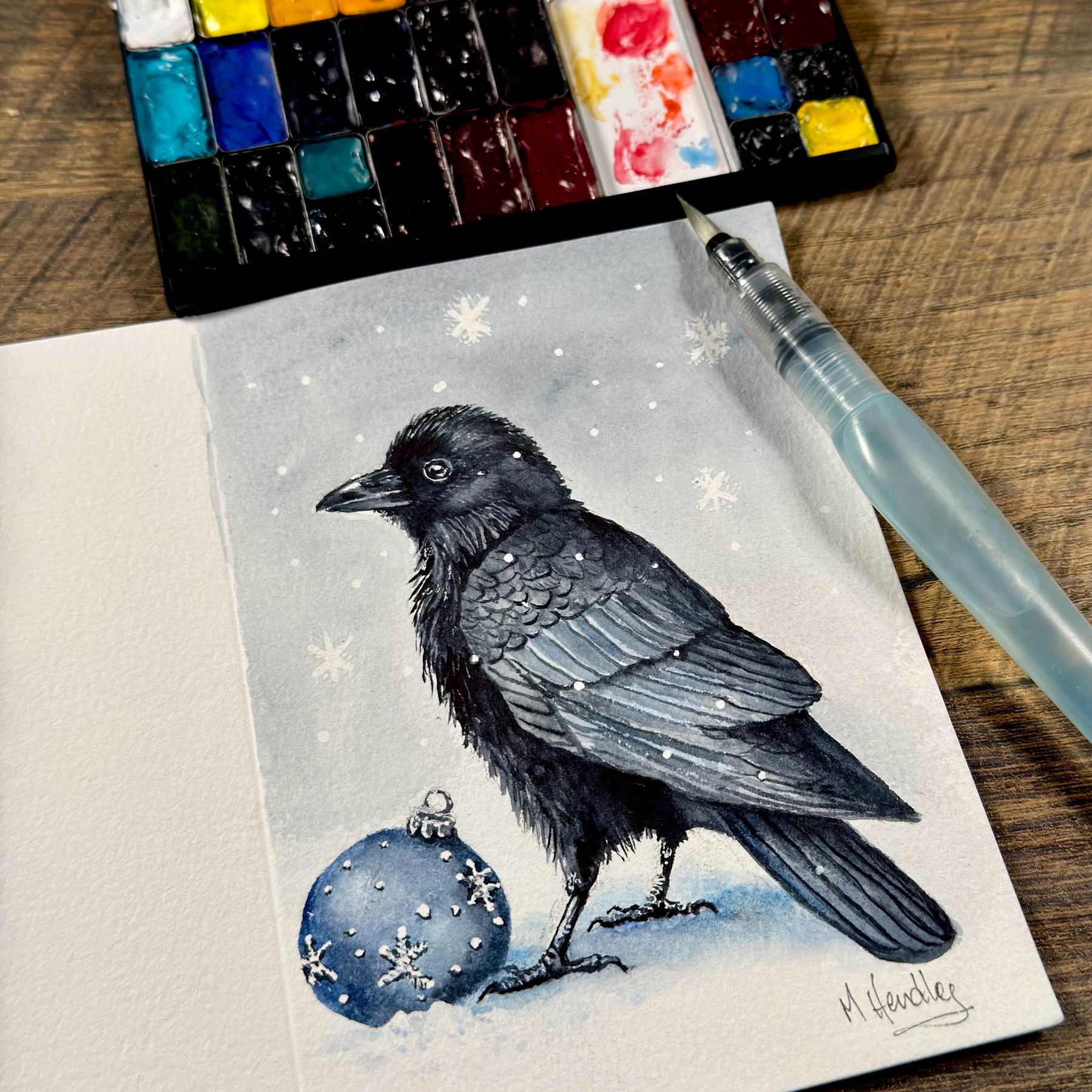 A watercolor painting of a crow standing in snow beside a blue ornament decorated with snowflakes. Snowflakes fall in the background. The painting is on a cold-press greeting card, shown alongside a watercolor palette and a Pentel water brush.&10;