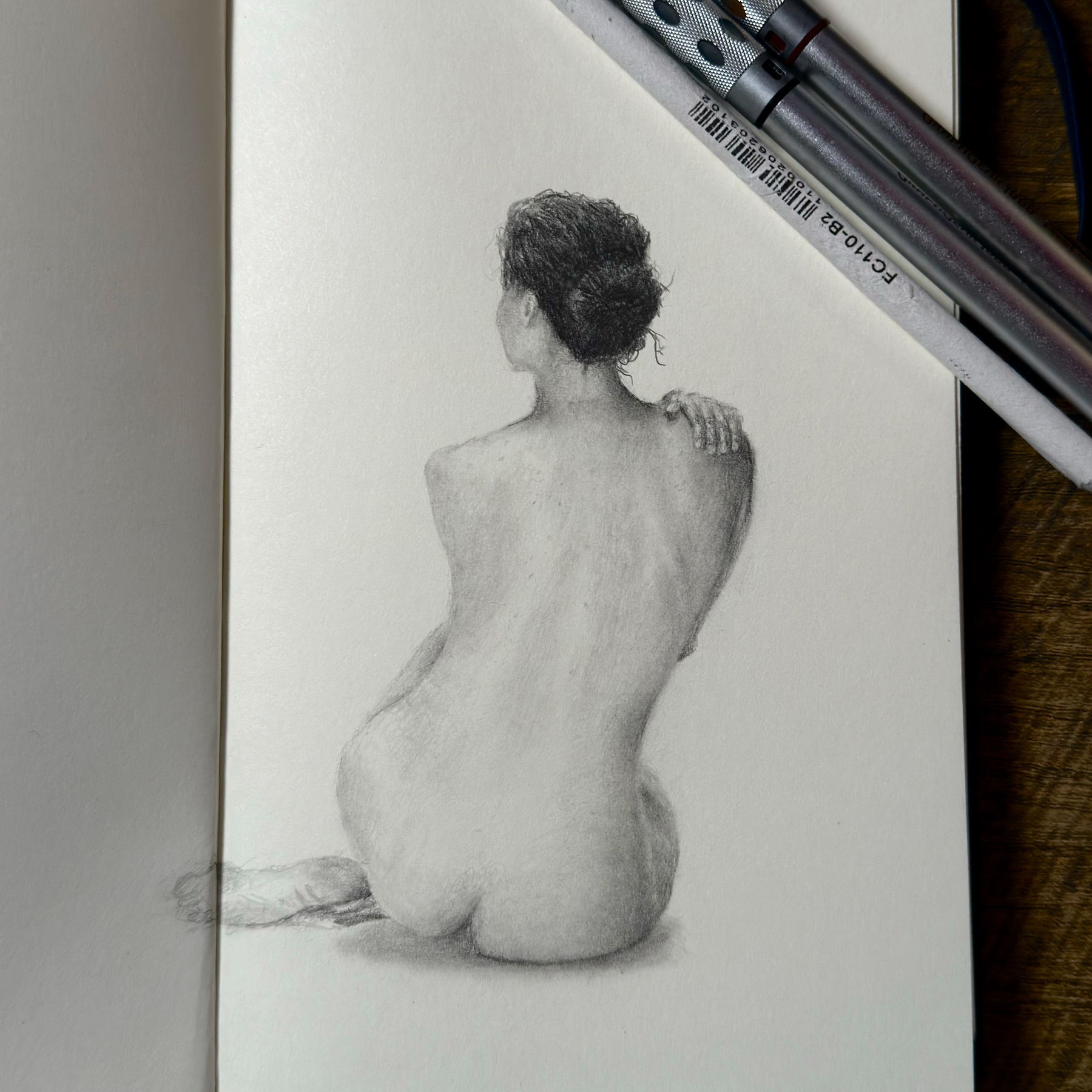 A pencil drawing of a woman's back, showing her seated with her knees pulled up, one hand resting on her shoulder. The focus is on the play of light and shadow over the curves of her back, hips, and bun-styled hair. Art materials are visible at the top right, suggesting an artist's workspace.