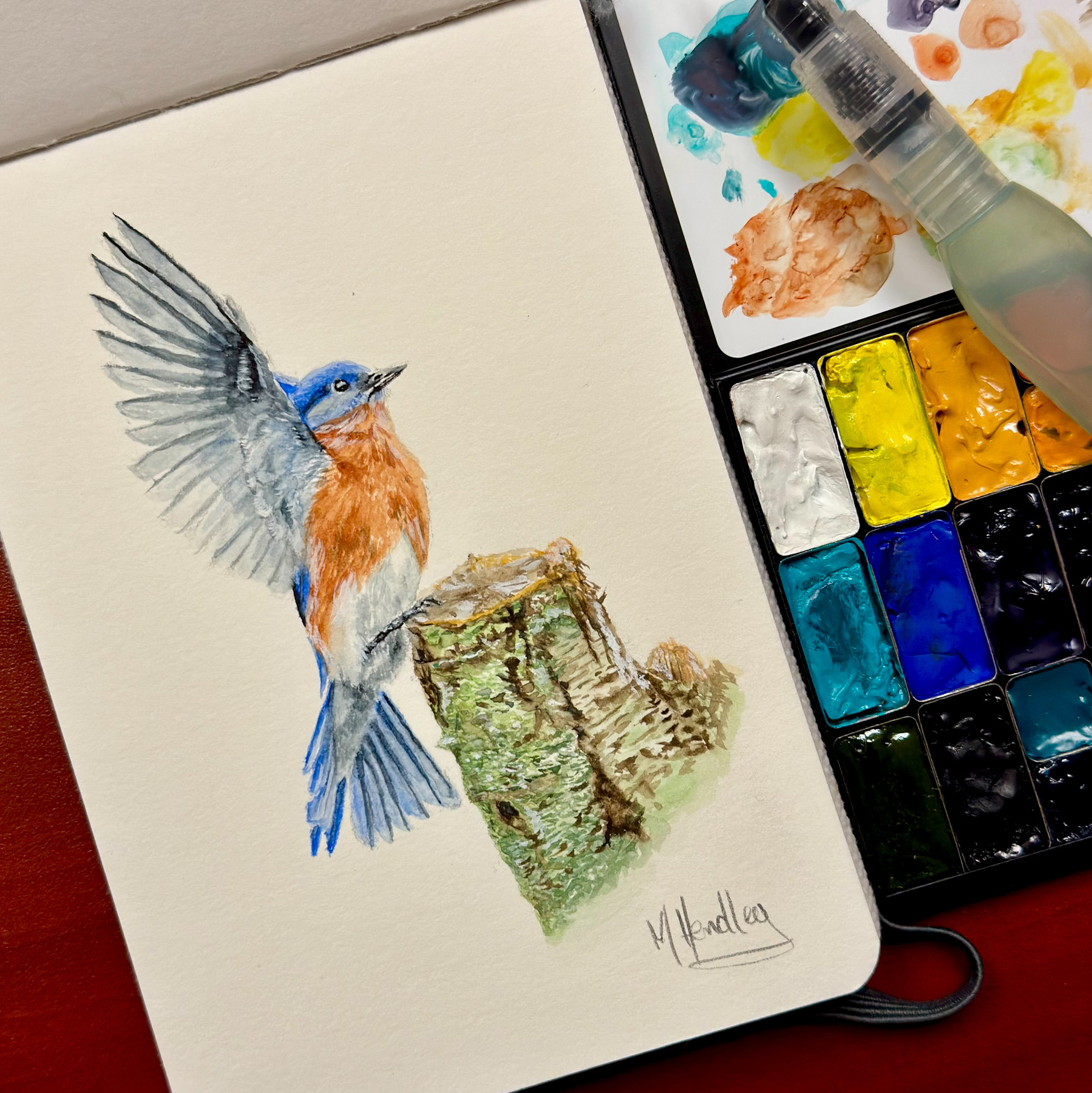 A watercolor painting of an eastern bluebird perched on a weathered birch stump, created with vibrant colors and fine details. A palette of QoR watercolors and a Pentel Aquash water brush are visible next to the artwork.&10;