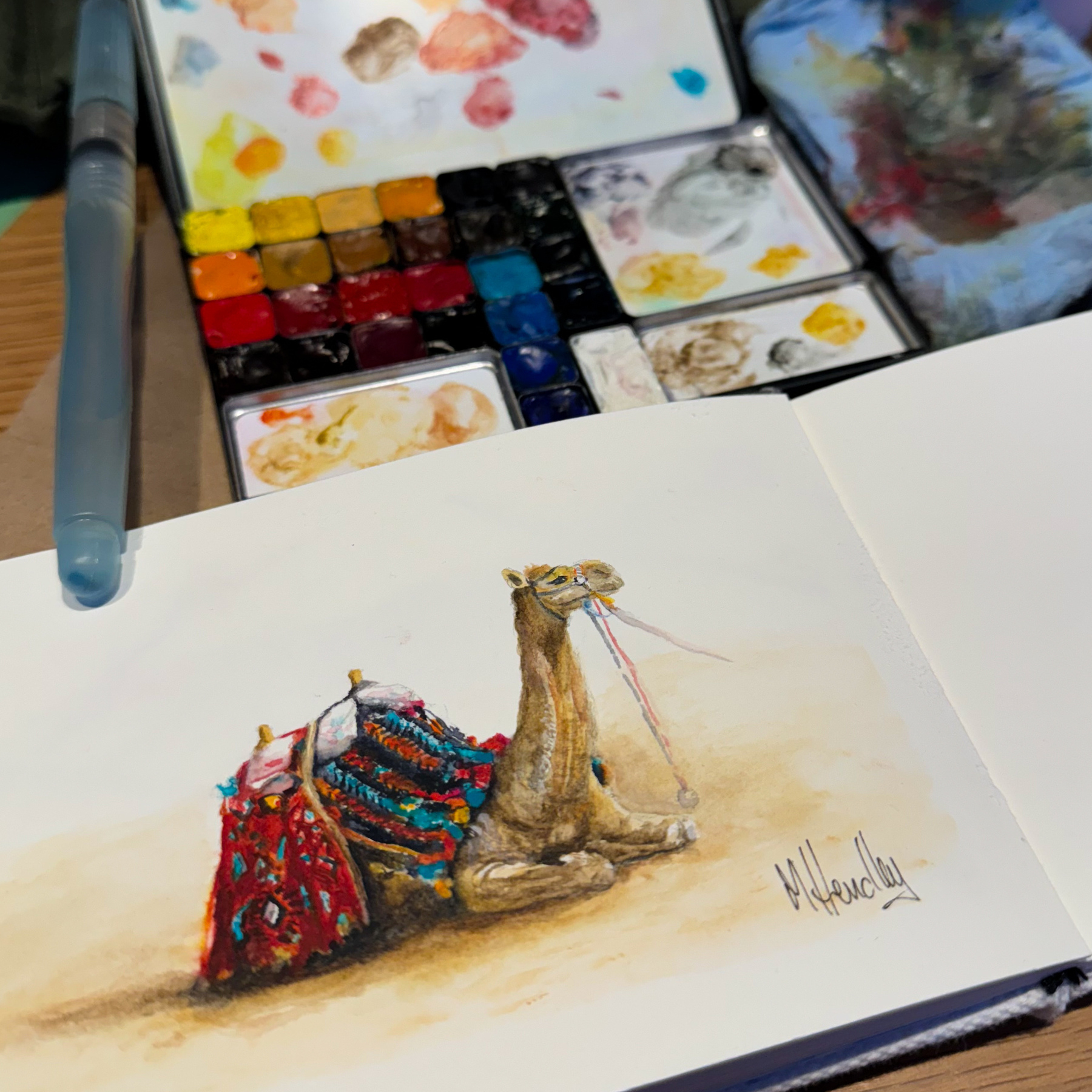 A watercolor painting of a camel adorned with colorful, intricate saddlery resting on a sandy ground is displayed next to an open watercolor palette with various colors. A water brush pen rests above the palette. The artist's signature is visible at the bottom of the painting. The palette and painting are on a wooden surface, suggesting an artist's workspace.