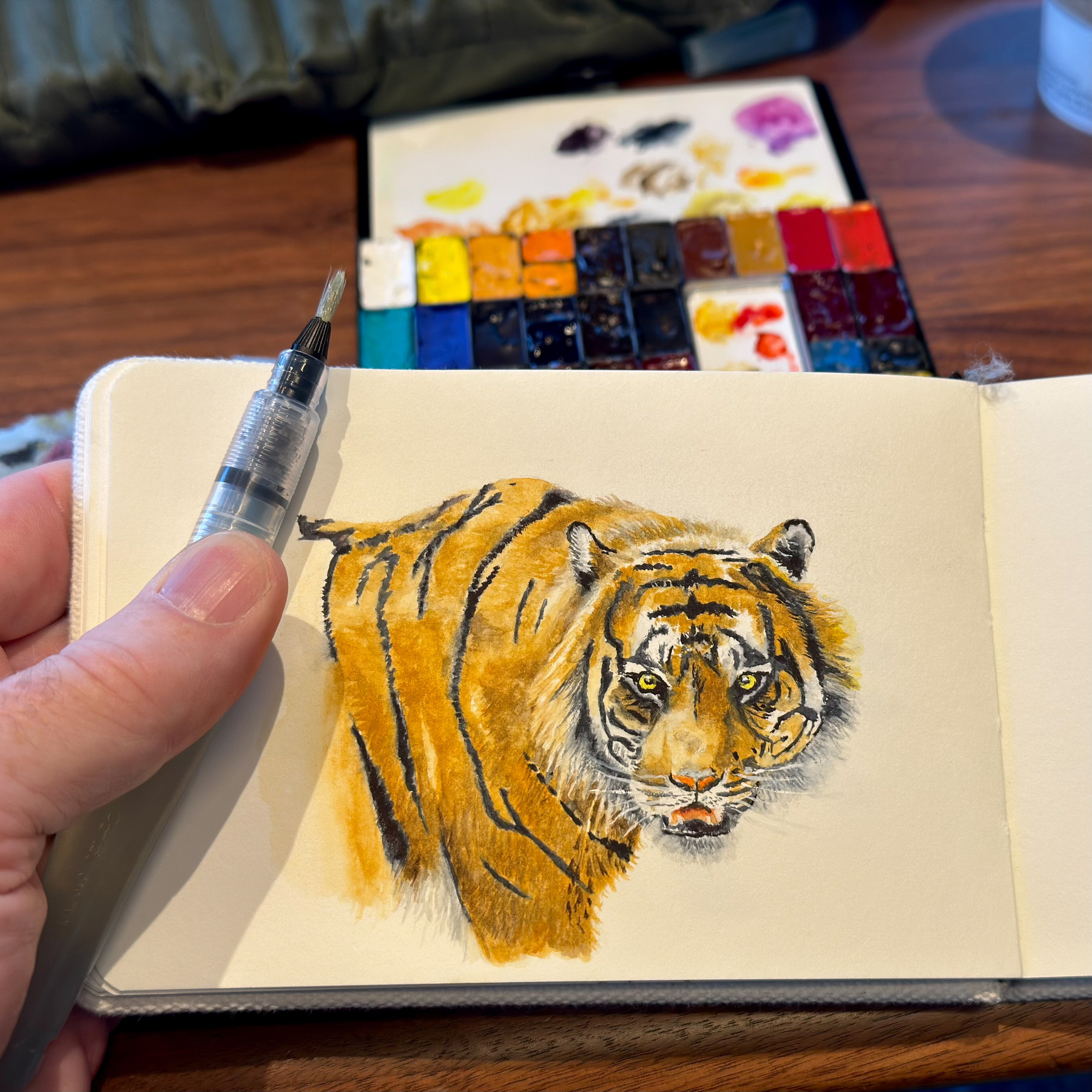 A close-up watercolor painting of a tiger in an Etchr A6 sketchbook, with vibrant orange and black stripes. The artist’s hand is visible holding a Pentel water brush, and a watercolor palette with various colors is seen in the background.