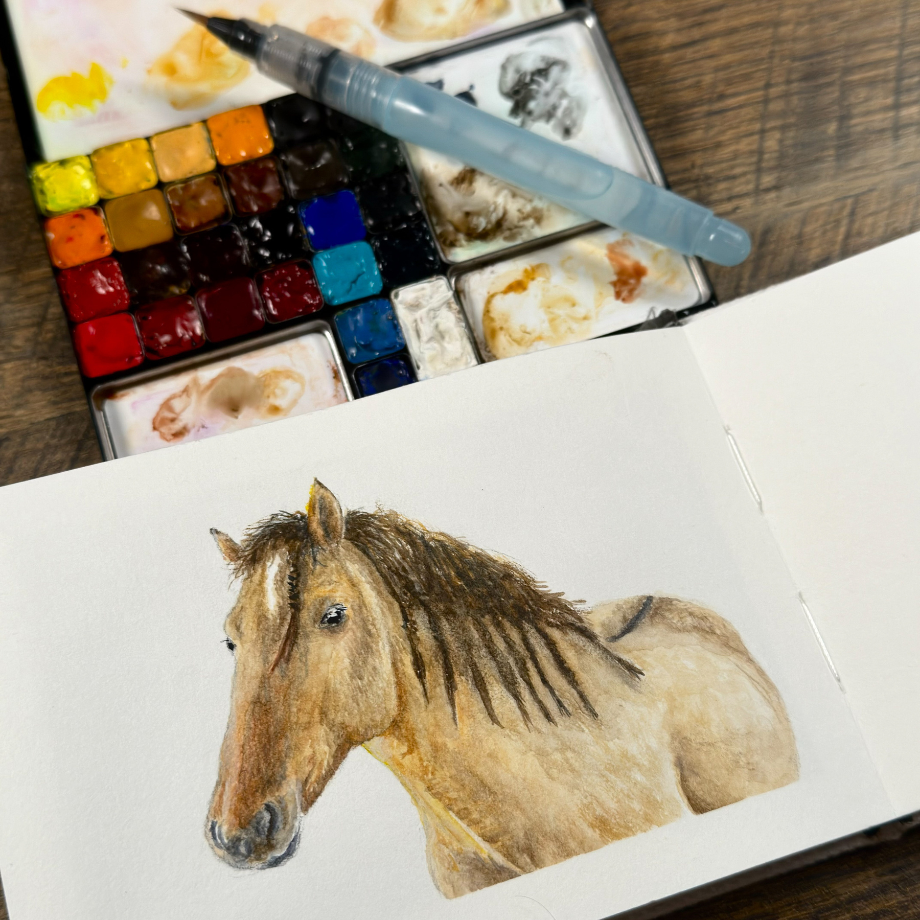 Watercolor painting of a horse on a sketchbook page, with a palette and water brush pen resting above.