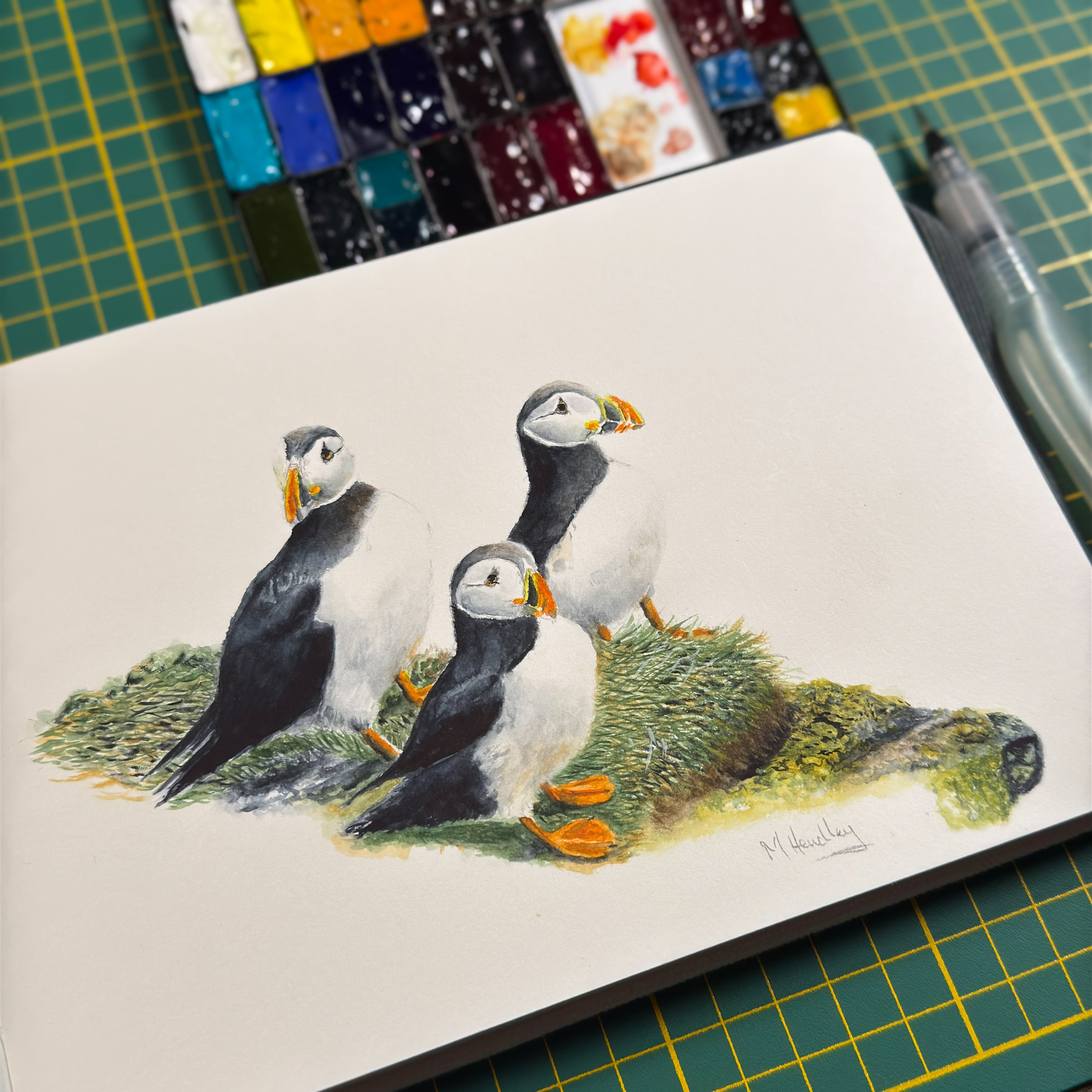 A watercolor painting of three puffins standing on grass, captured in progress. The painting is in an open A5 sketchbook, placed on a wooden table. Next to the sketchbook are a palette with vibrant watercolor paints and various drawing tools, including a water brush.&10;