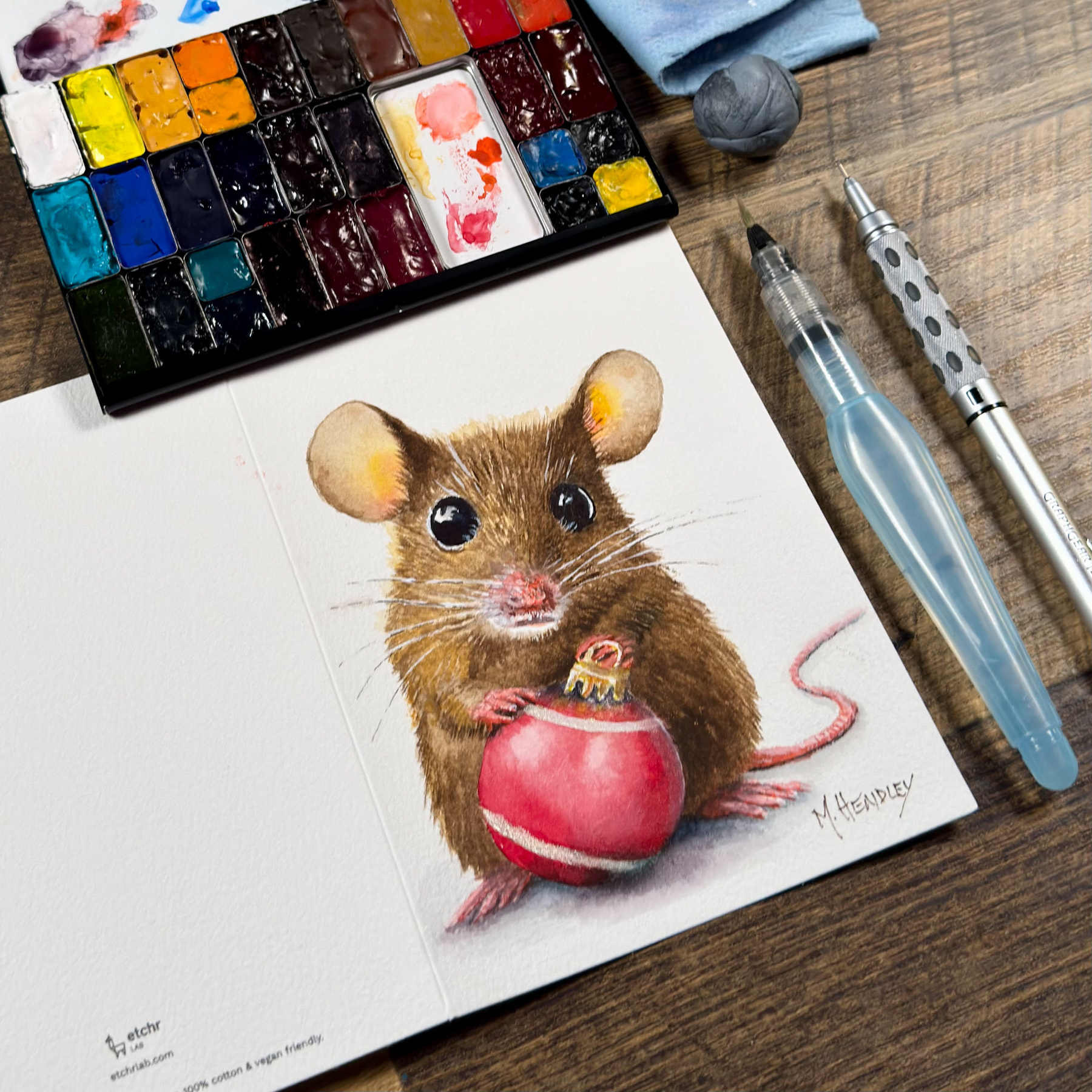 A detailed watercolor painting of a brown mouse holding a red Christmas ornament, created on a folded Etchr cold press greeting card. Nearby are a watercolor palette, a Pentel aquash brush pen, and a mechanical pencil on a wooden surface.