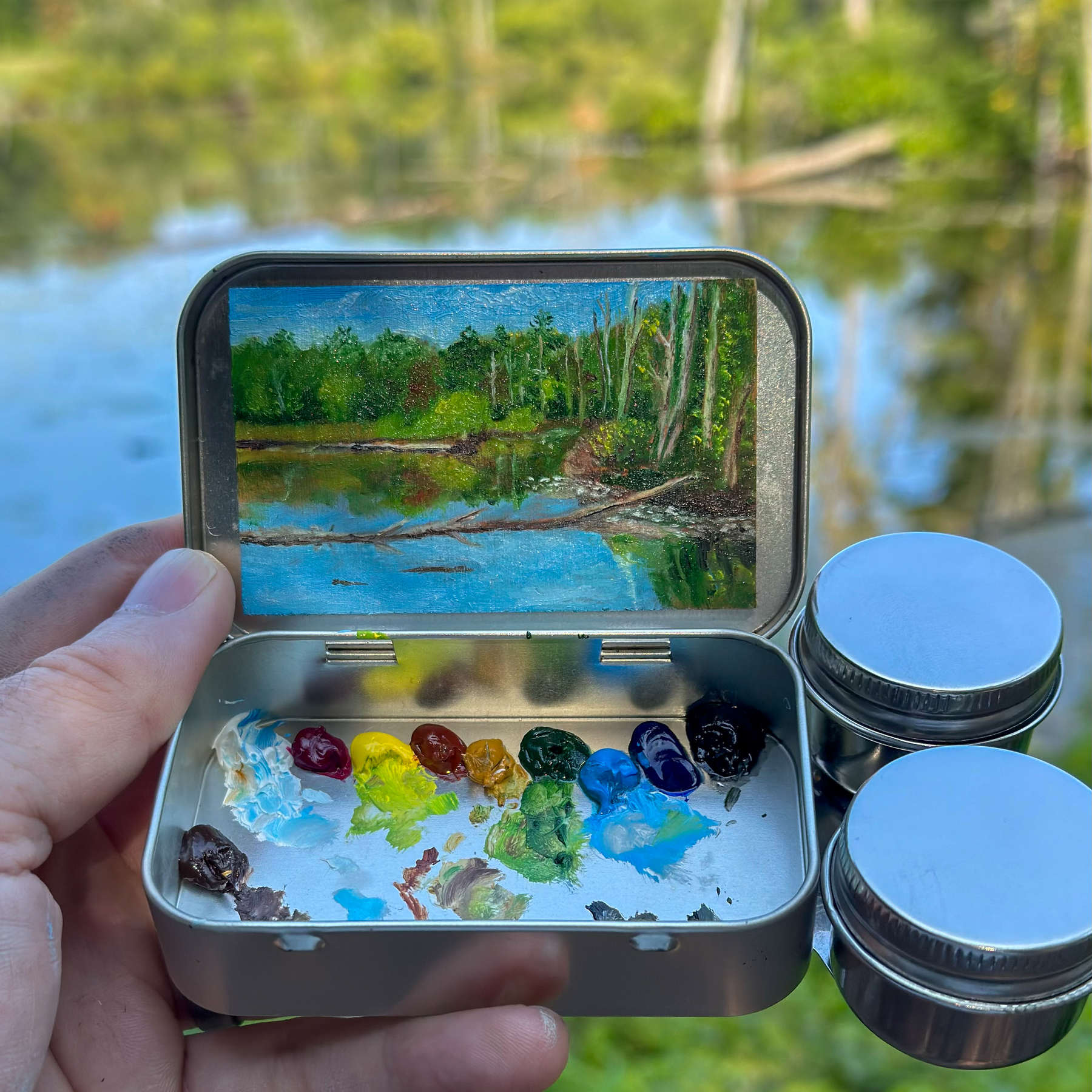 A small landscape painting inside an open Altoid tin, depicting a tranquil lake surrounded by lush green trees. The painting is done with oil paints, which are visible in the bottom half of the tin, arranged in a colorful palette. The real-life scene of the lake is blurred in the background.