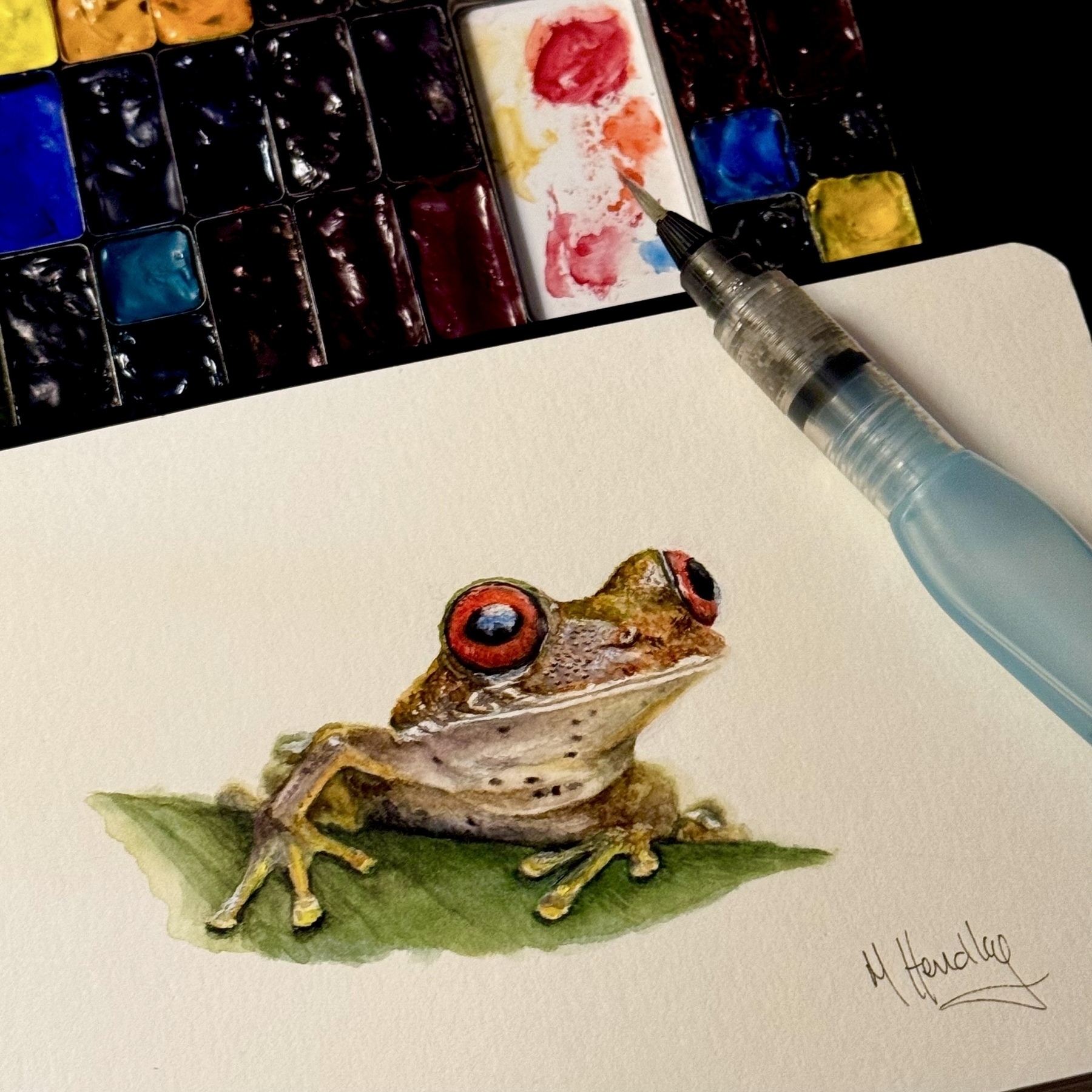 A detailed painting of a tree frog on a leaf is accompanied by a watercolor set and brush.