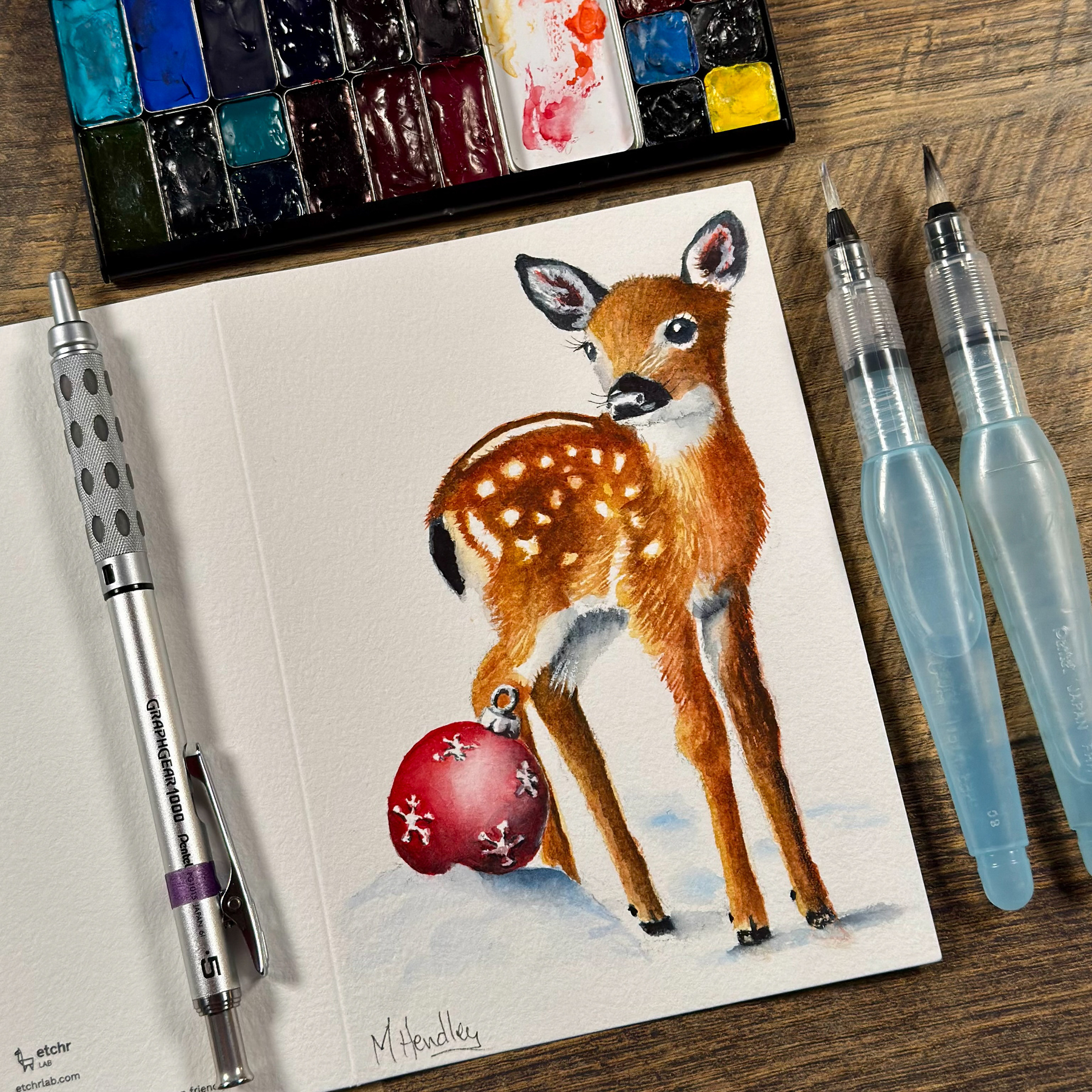 A watercolor painting on a greeting card of a fawn standing on snow, with a red ornament featuring white snowflake patterns resting near its back leg. Tools and materials like water brushes, a mechanical pencil, and a watercolor palette are visible around the card.