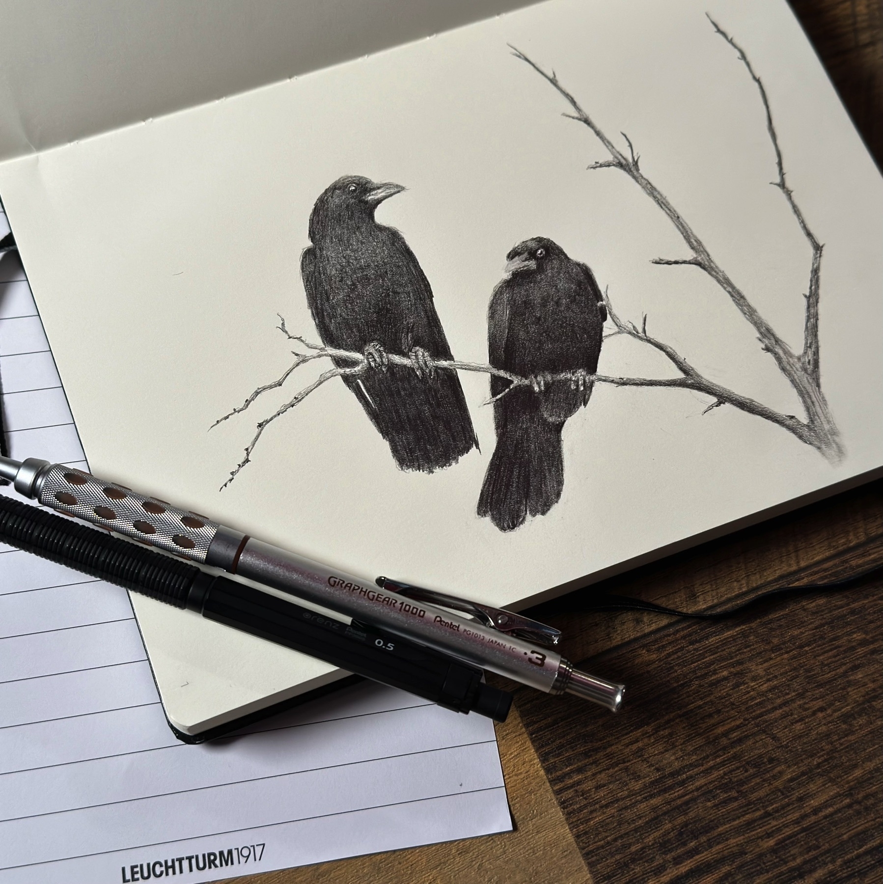 A pencil drawing of two crows perched on a branch, with the drawing situated on an open sketchbook page. A mechanical pencil rests on the sketchbook's edge, next to the drawing. The sketchbook is placed on a wooden table, partially atop a piece of paper with grid lines. The image conveys a calm, studious atmosphere often associated with an artist's workspace.